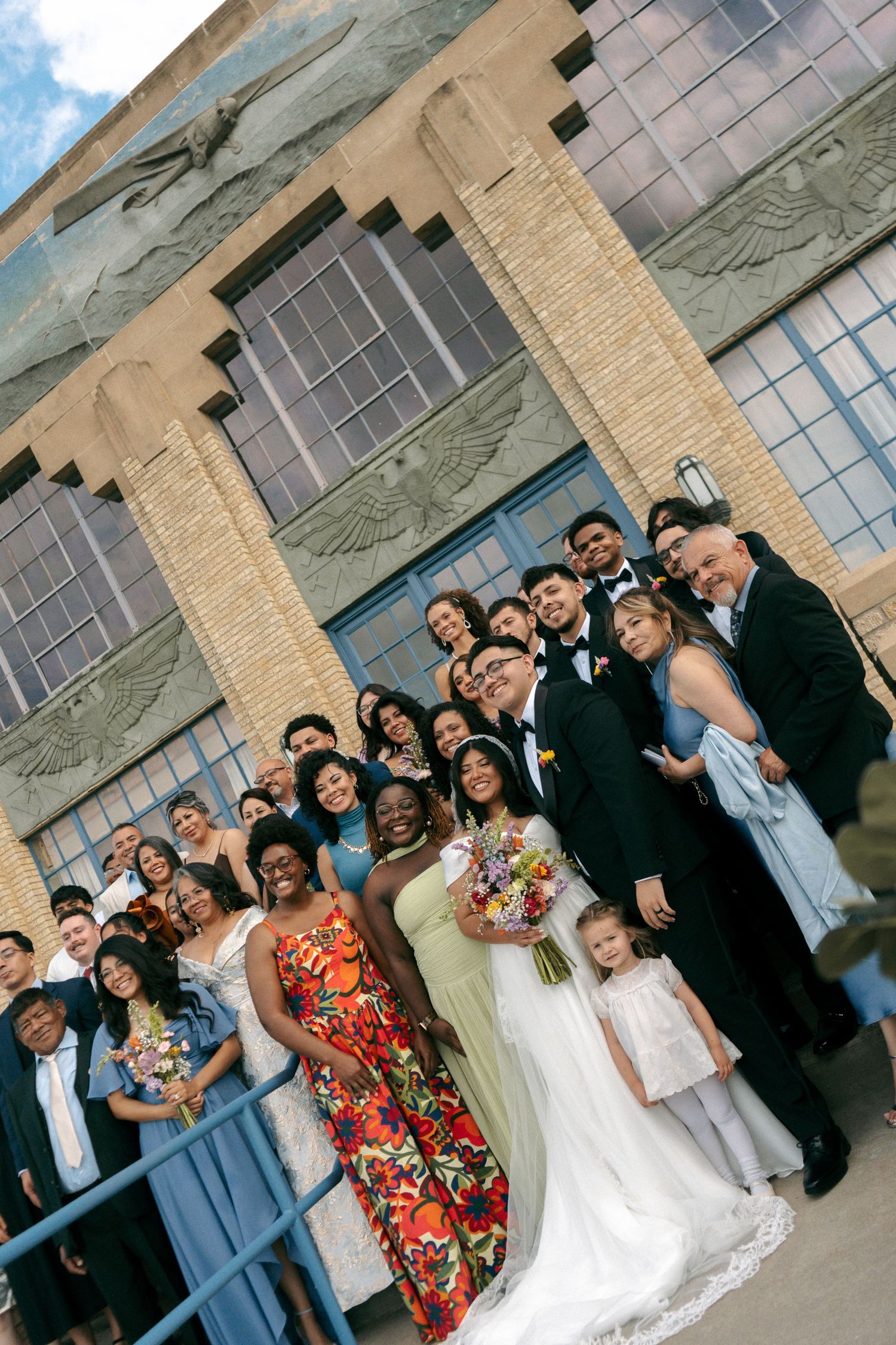 Wedding party poses outside a brick building, bride holding colorful bouquet surrounded by guests in formal attire.