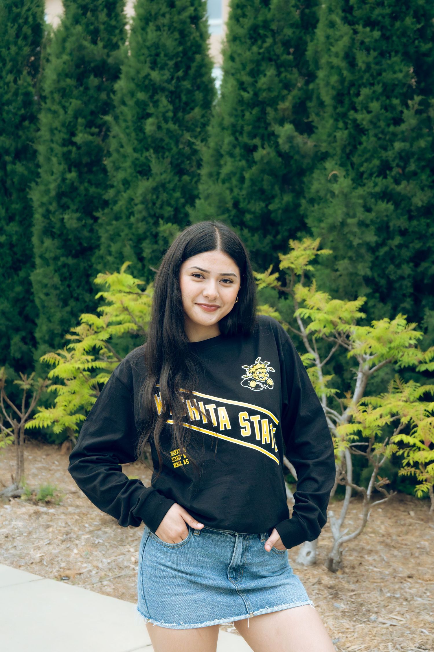 Young woman in black Wichita State sweatshirt and denim skirt posing near green trees.