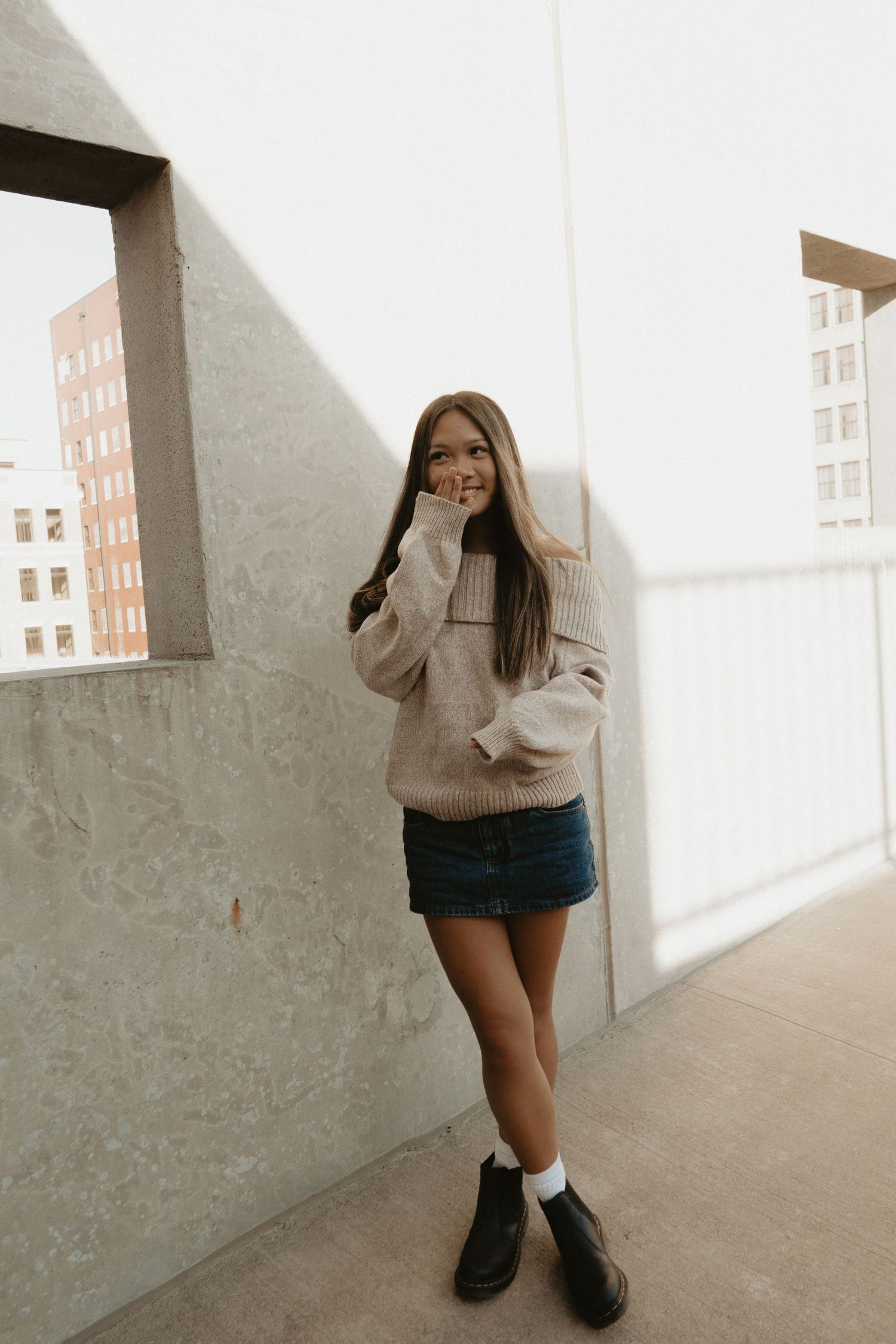 Young woman smiling in beige sweater and denim mini skirt leaning against concrete wall on urban rooftop.