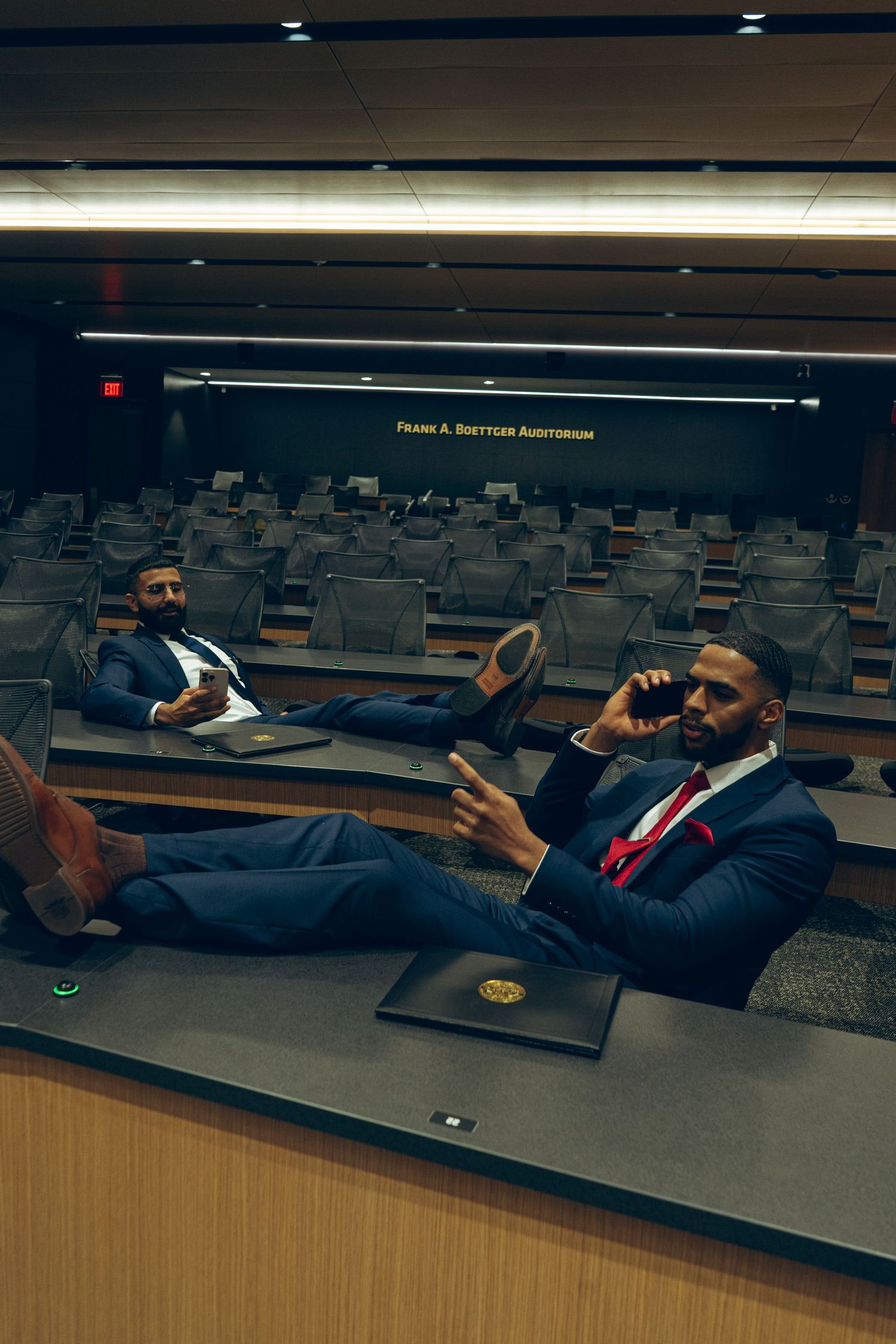 Two businessmen in navy suits talking on phones while relaxing in an empty auditorium.