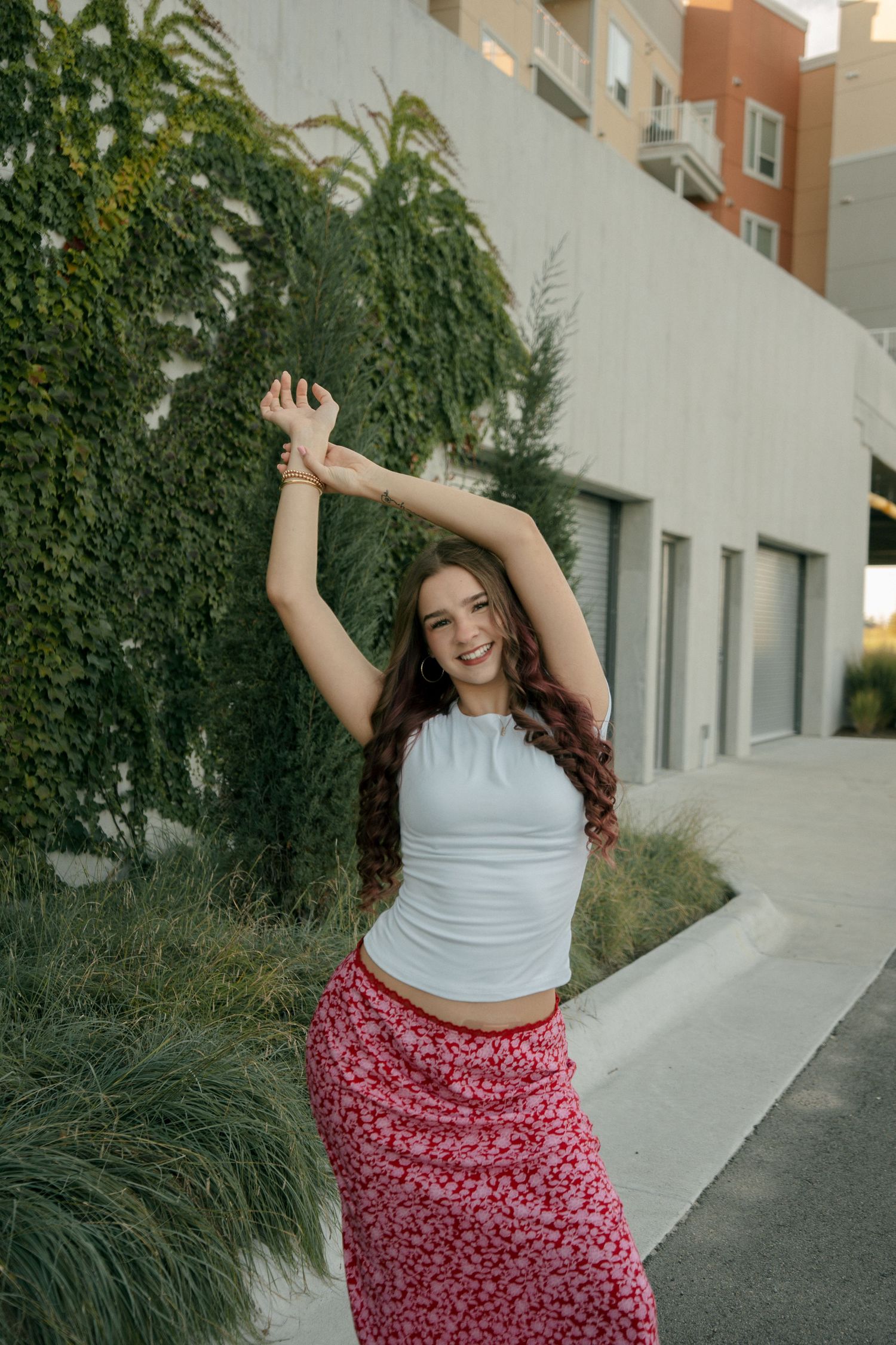Young woman in white crop top and red floral skirt posing with arms raised outdoors near ivy-covered wall.