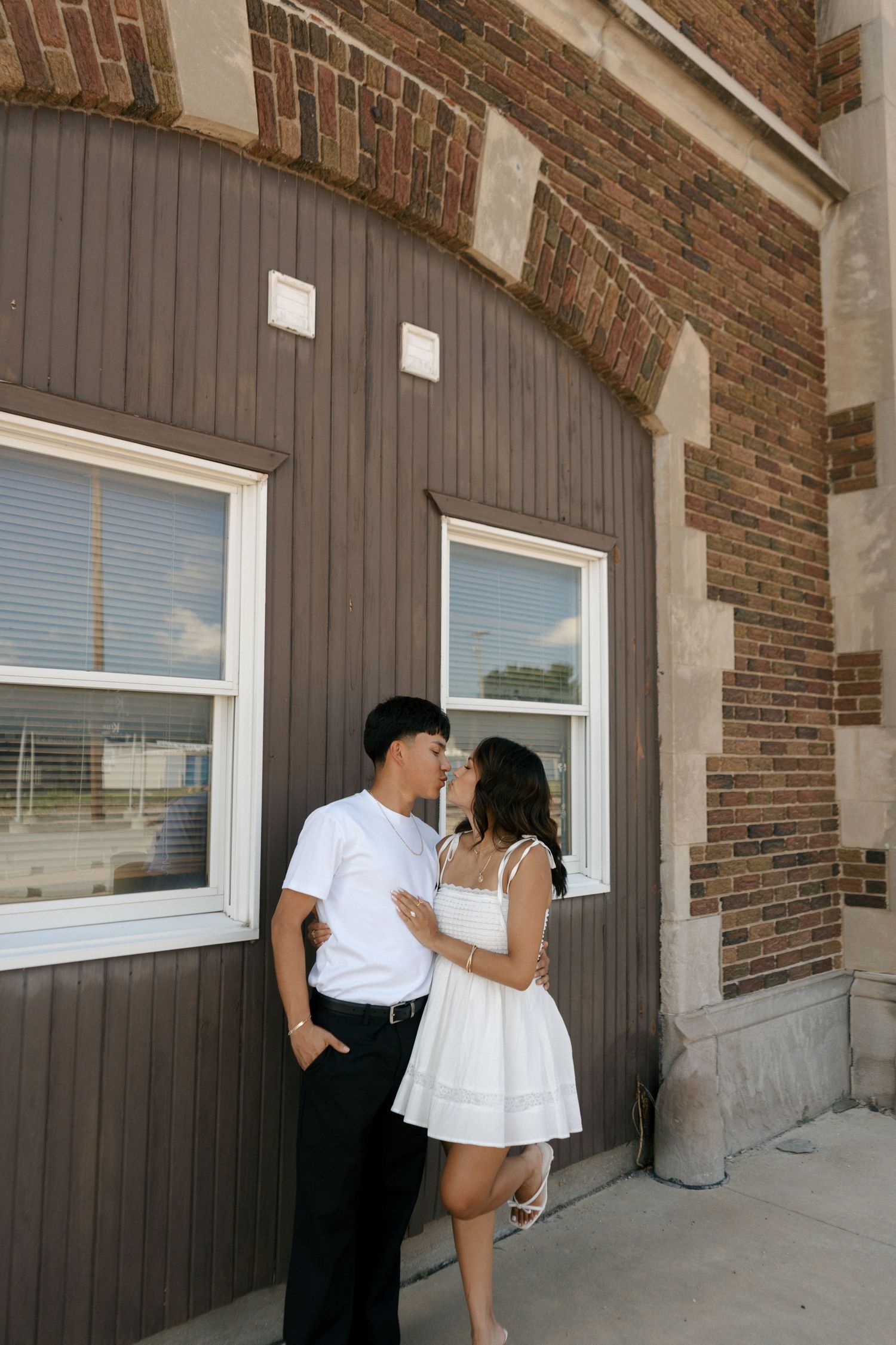 Couple sharing a romantic moment near a brick building arch, woman in white dress and man in white shirt.