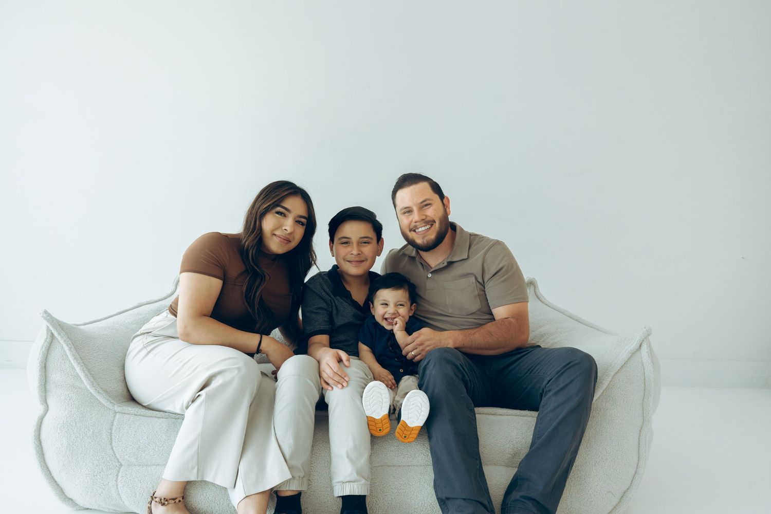 Happy family of four posing together on a white couch in a bright studio.