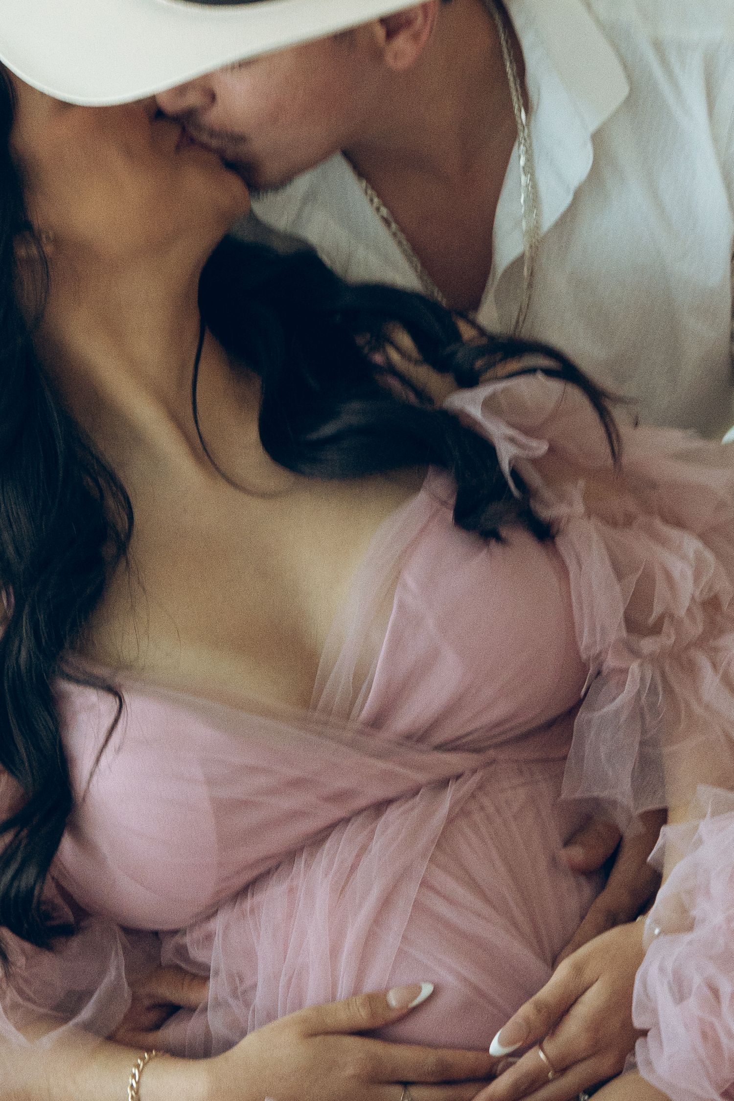 Couple sharing a kiss during maternity photoshoot, man in white cowboy hat embracing pregnant woman in pink tulle gown.