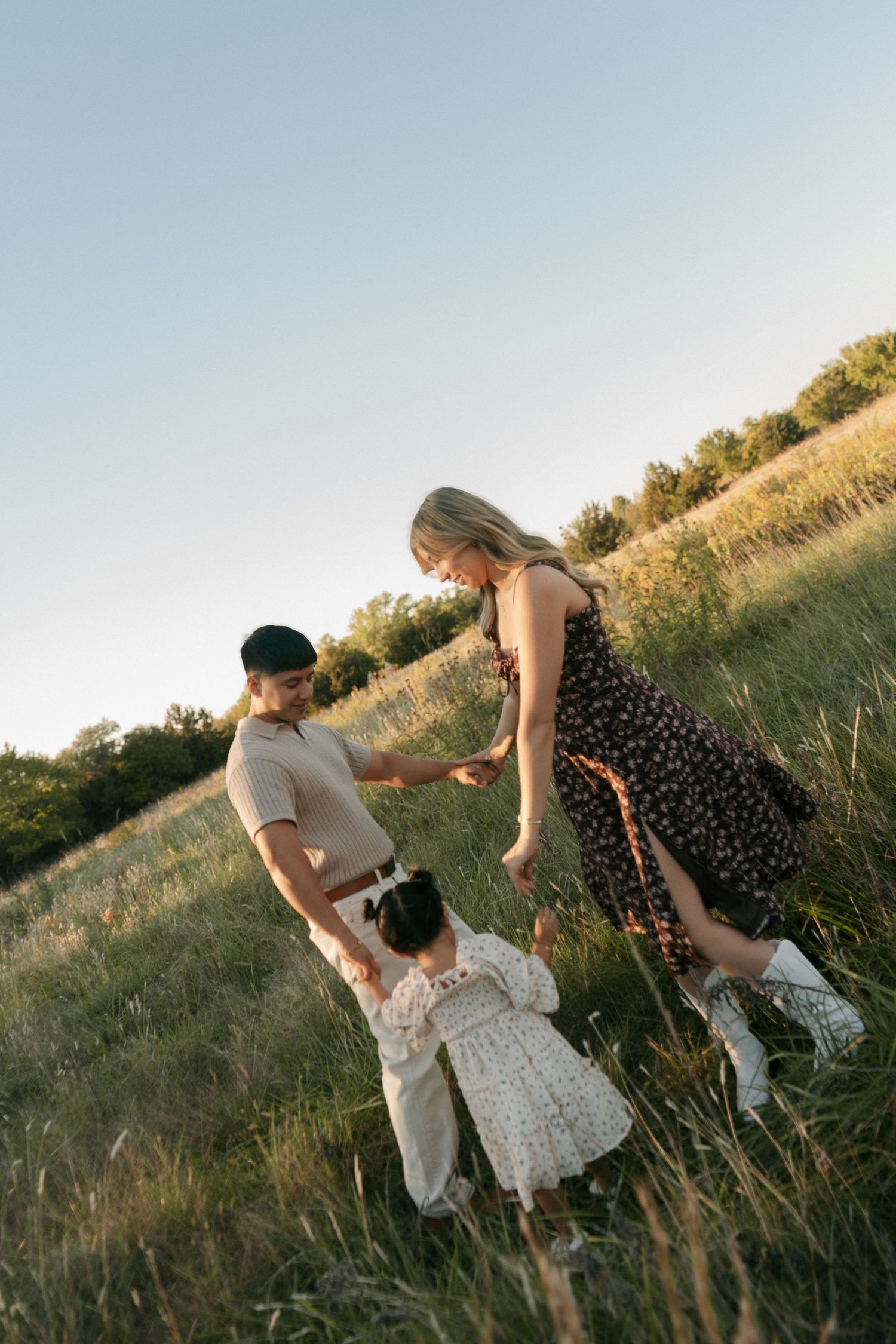 A couple and their young daughter hold hands and interact playfully on a grassy hillside during a warm sunset.