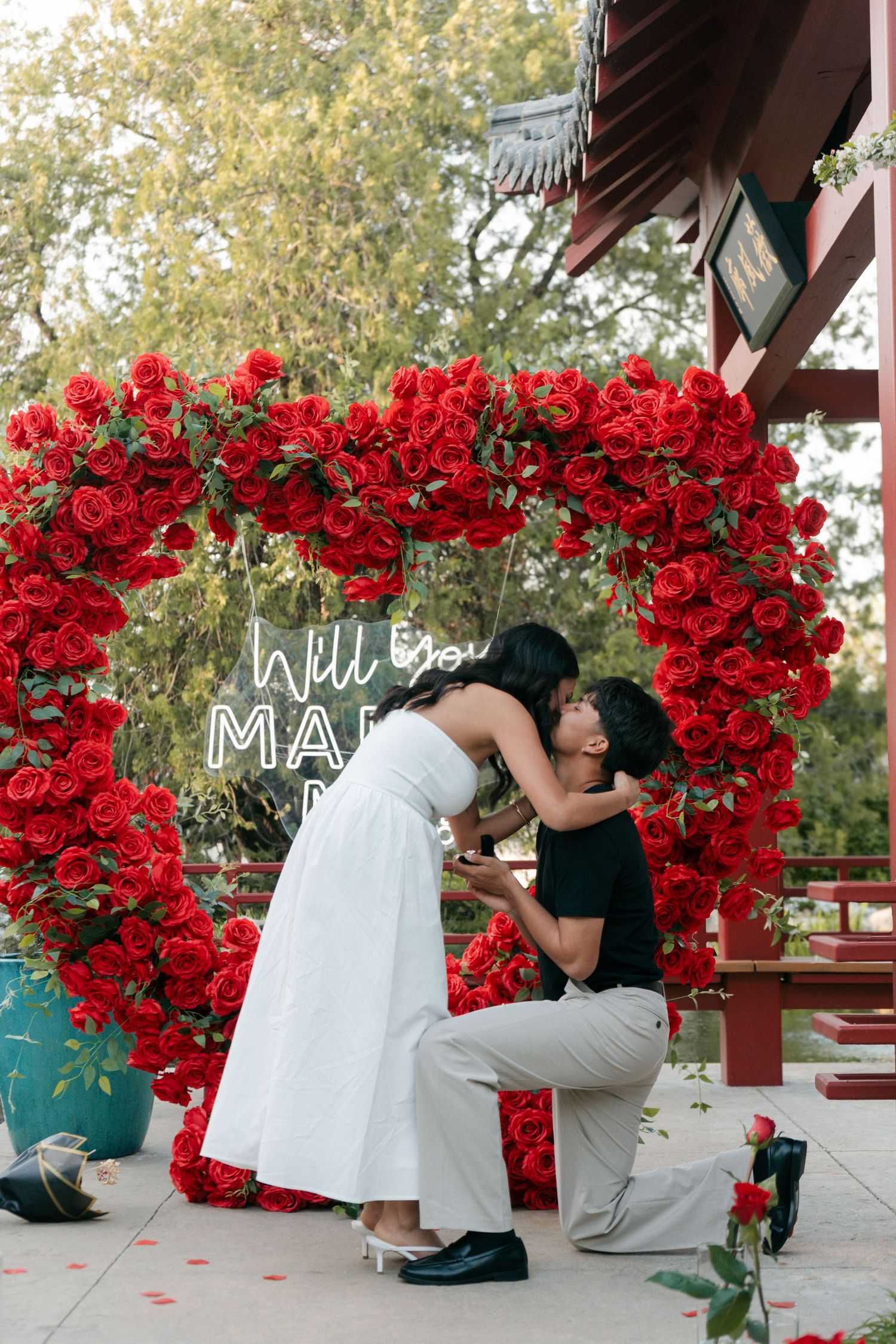 Newly engaged couple sharing a kiss under a red rose heart arch with Will You Marry Me neon sign.