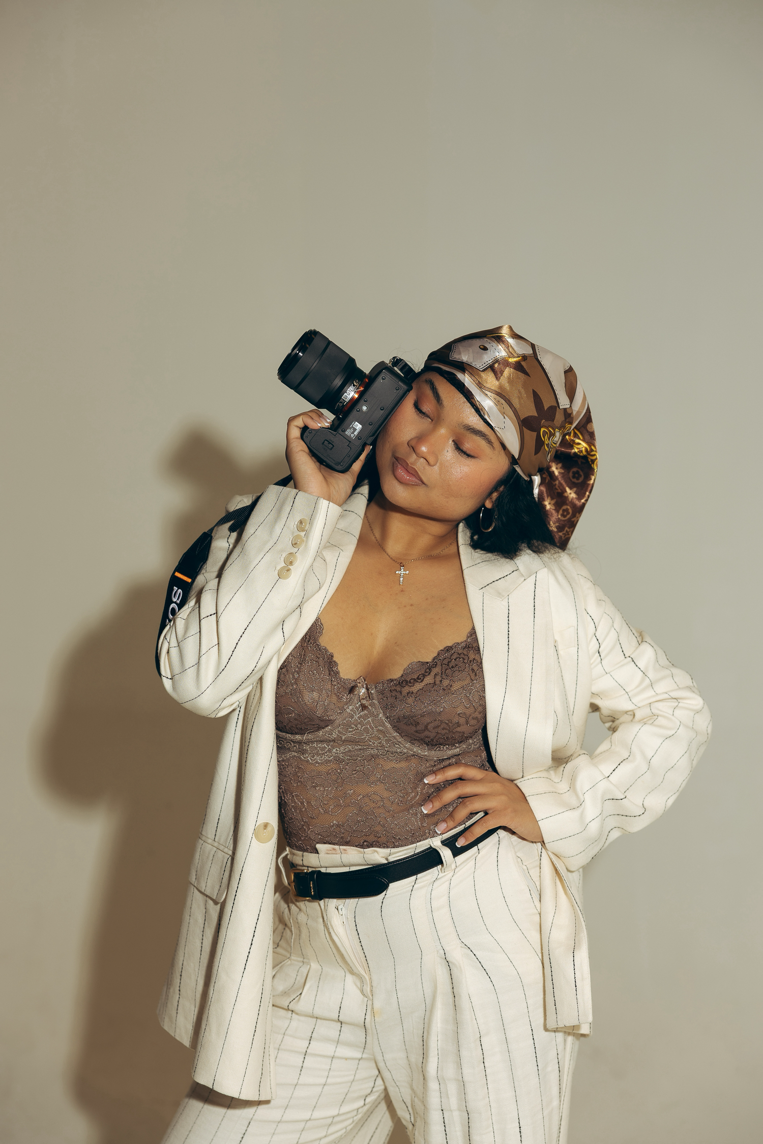 Young woman in white pinstripe suit holding camera, wearing lace top and head scarf, posing confidently.