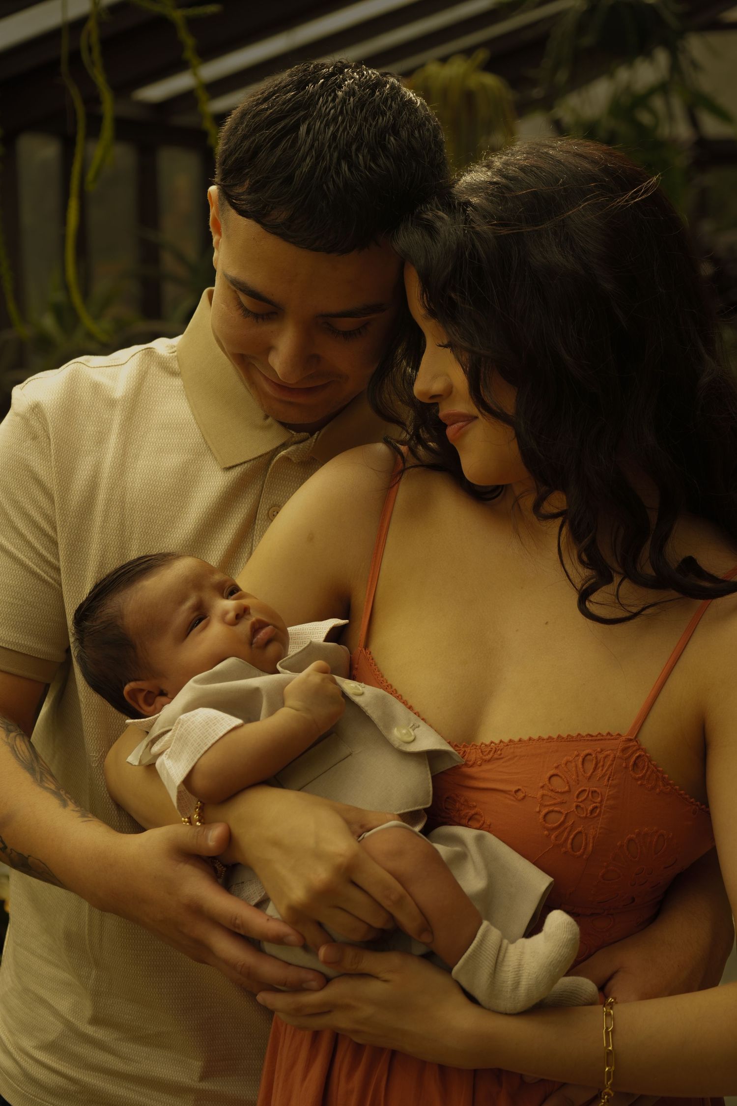 Young couple smiling down at newborn baby they cradle together in a lush greenhouse setting.