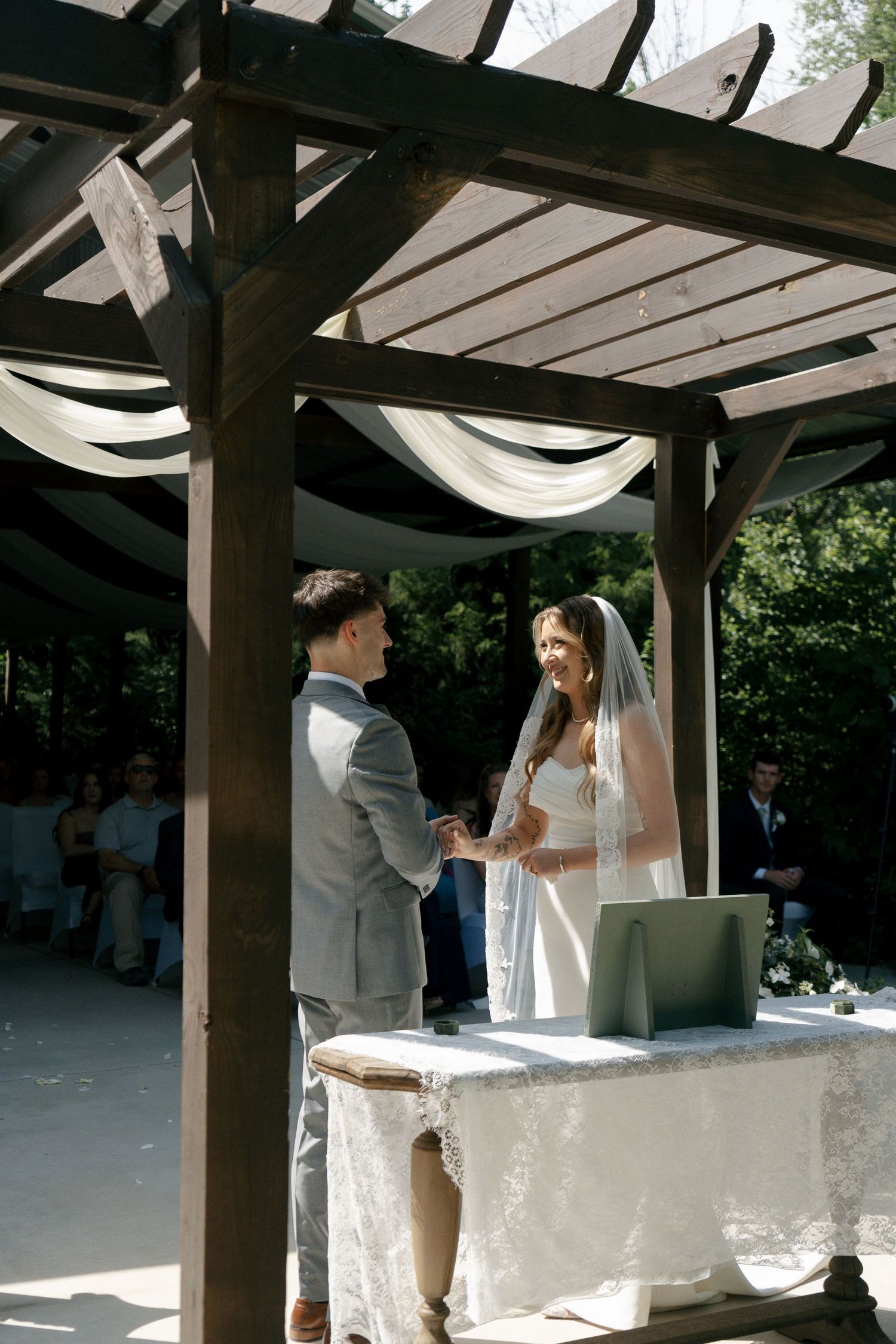 Bride and groom exchange vows under a wooden pergola decorated with white draping at an outdoor wedding ceremony.