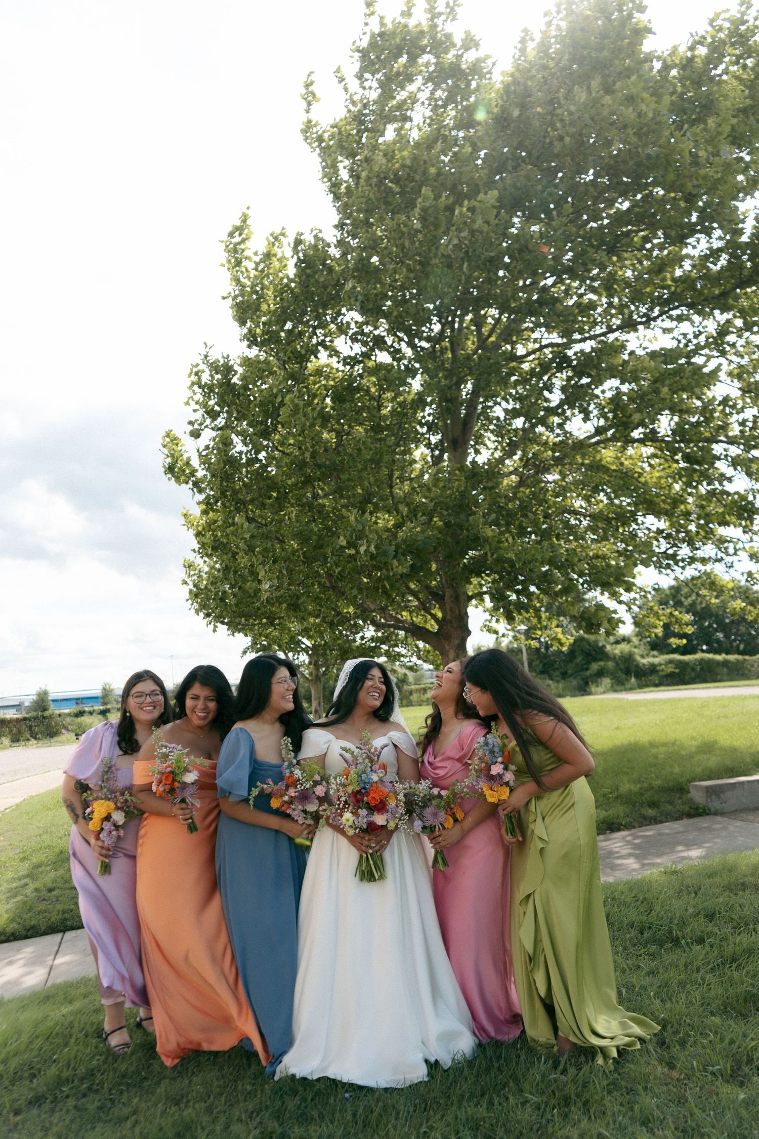 Bride in white gown poses with five bridesmaids in colorful mismatched dresses holding bouquets outdoors near a tree.