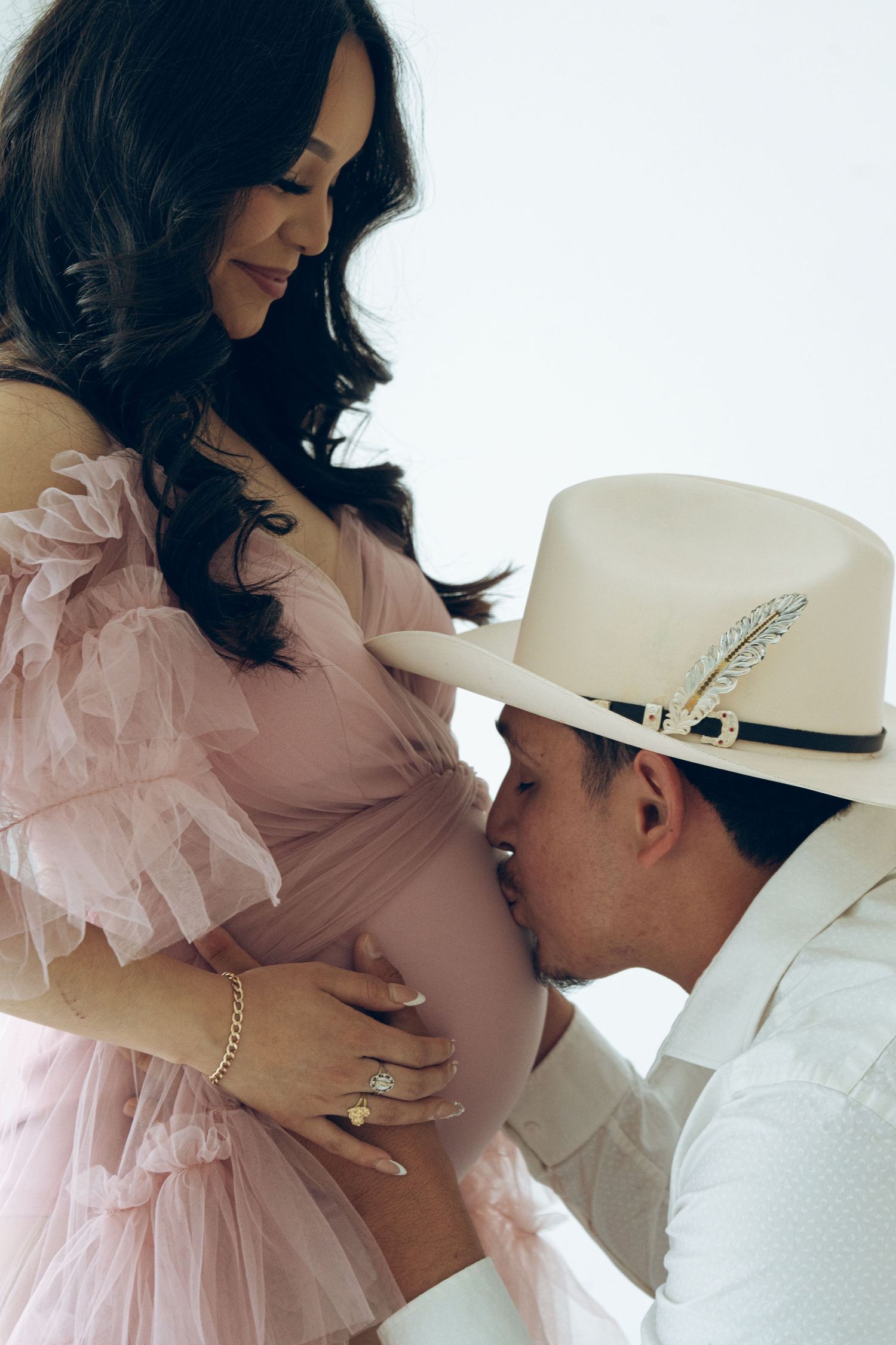 Man in white cowboy hat kisses pregnant partners baby bump during maternity photoshoot in white studio.