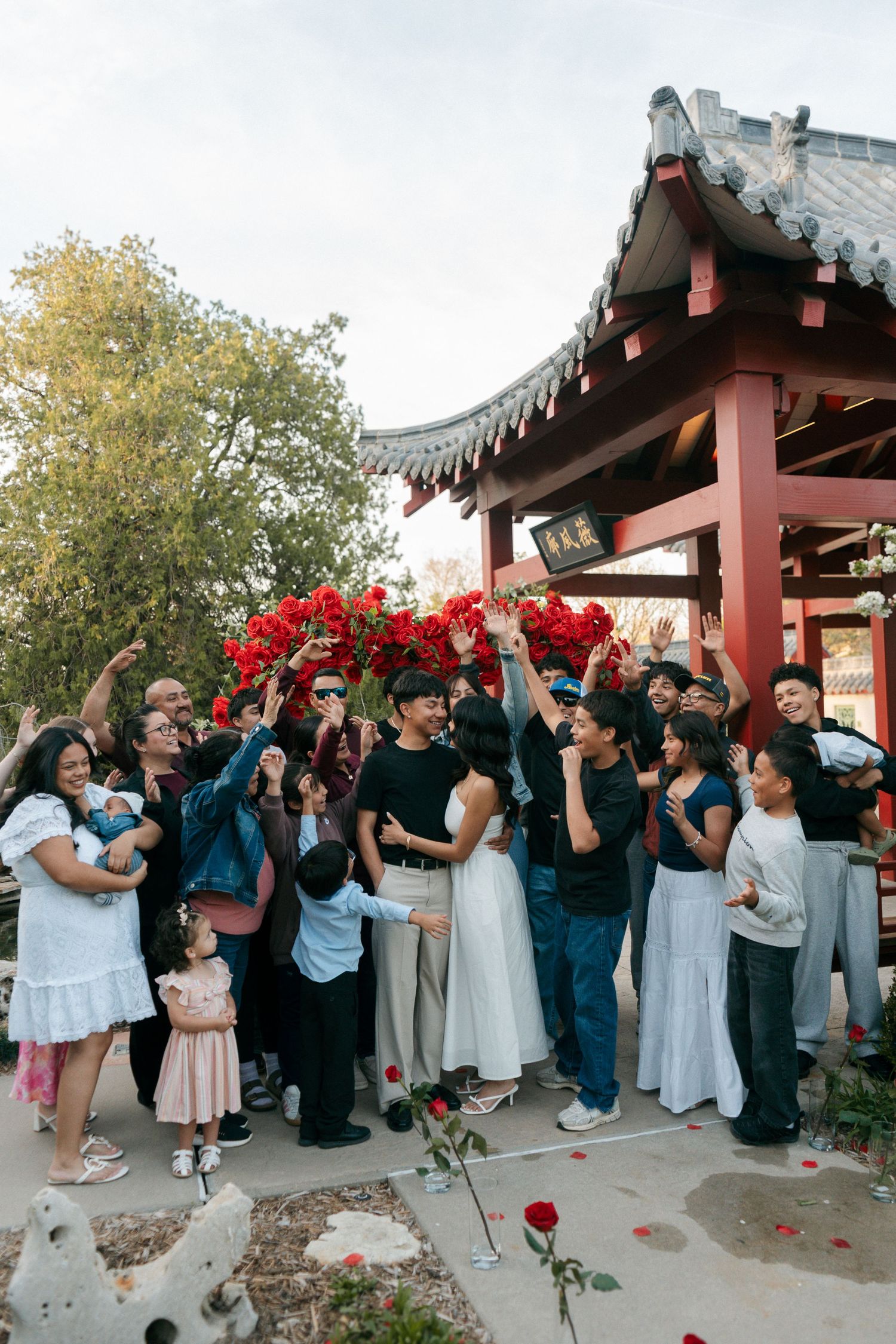 Joyful group of people celebrating under a red flower arch at a Chinese garden pavilion.
