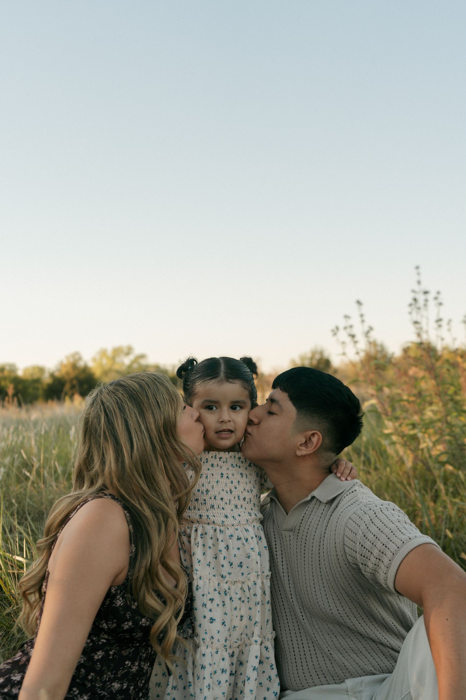 Two parents lovingly kiss their young daughter on the cheeks while sitting in a golden sunlit meadow.