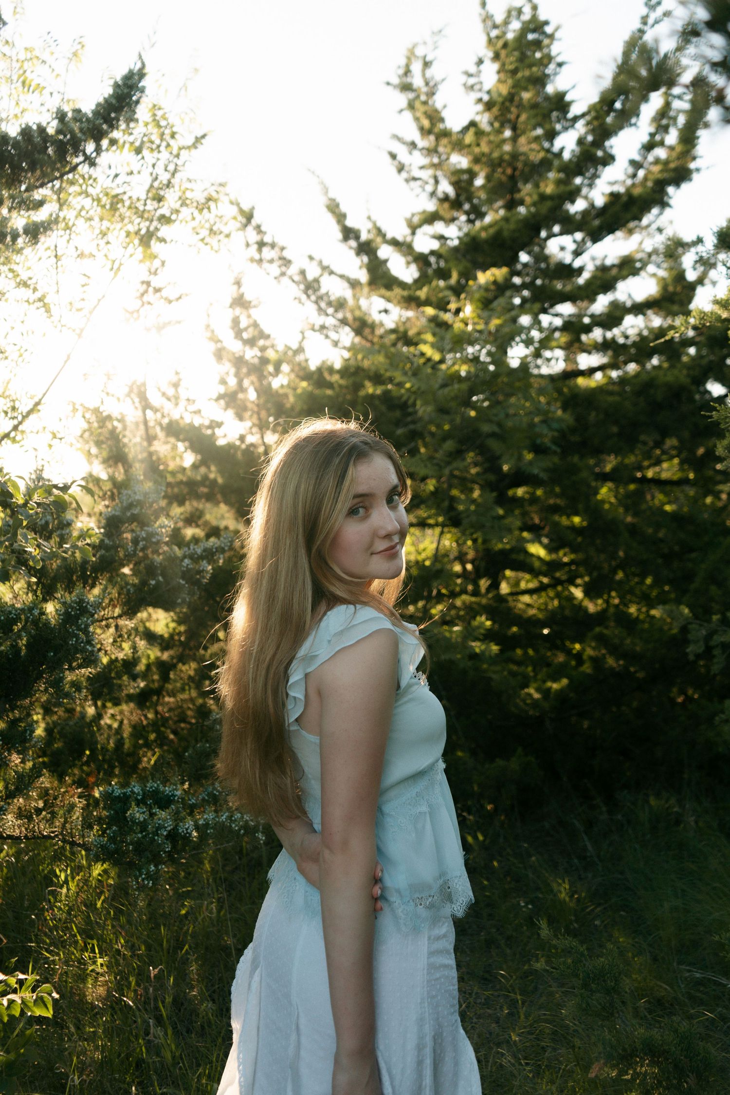 Young woman in white dress standing in sunlit forest clearing with evergreen trees glowing in golden hour light.