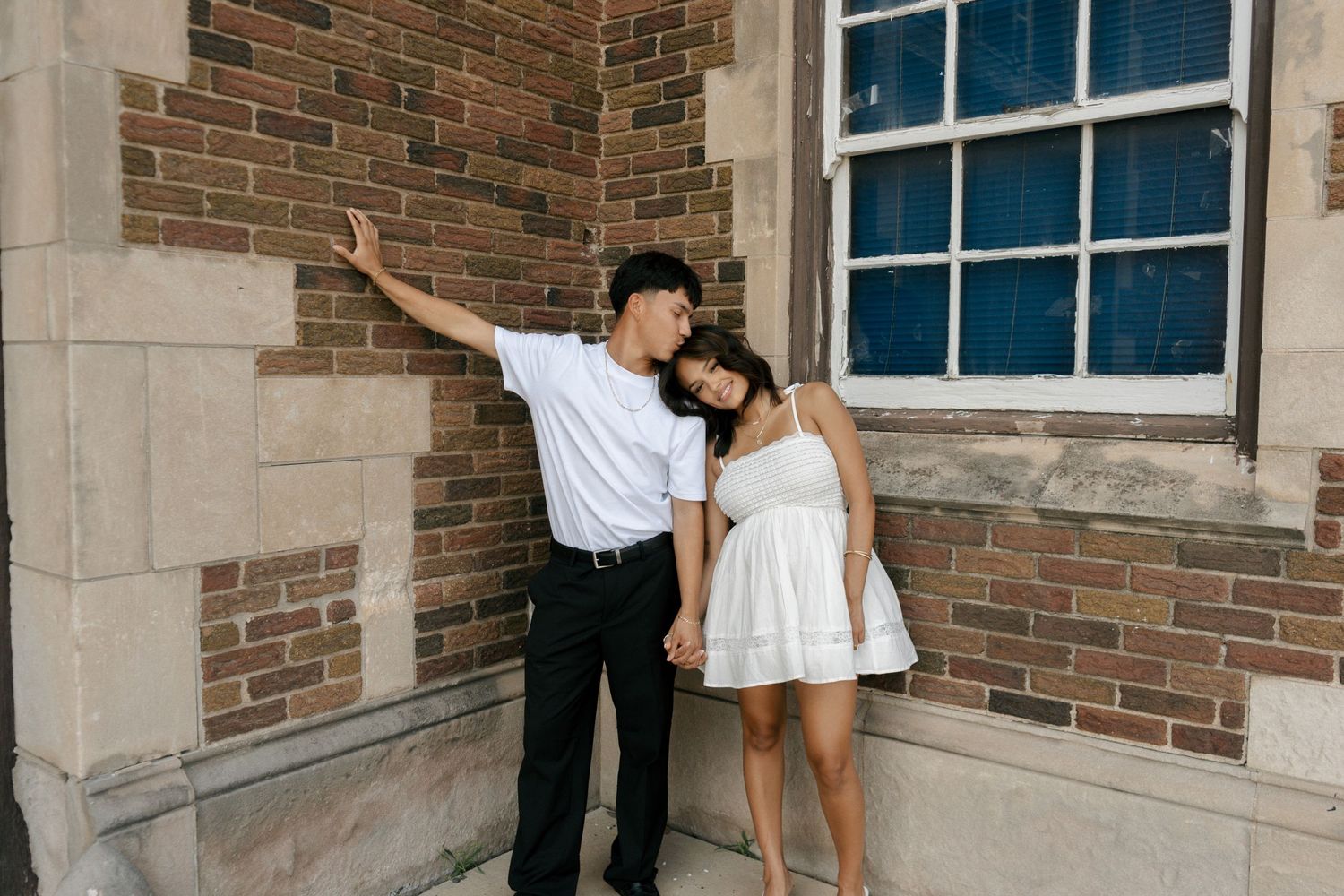Couple leaning against a brick building wall, woman in white dress, man in white shirt and black pants.