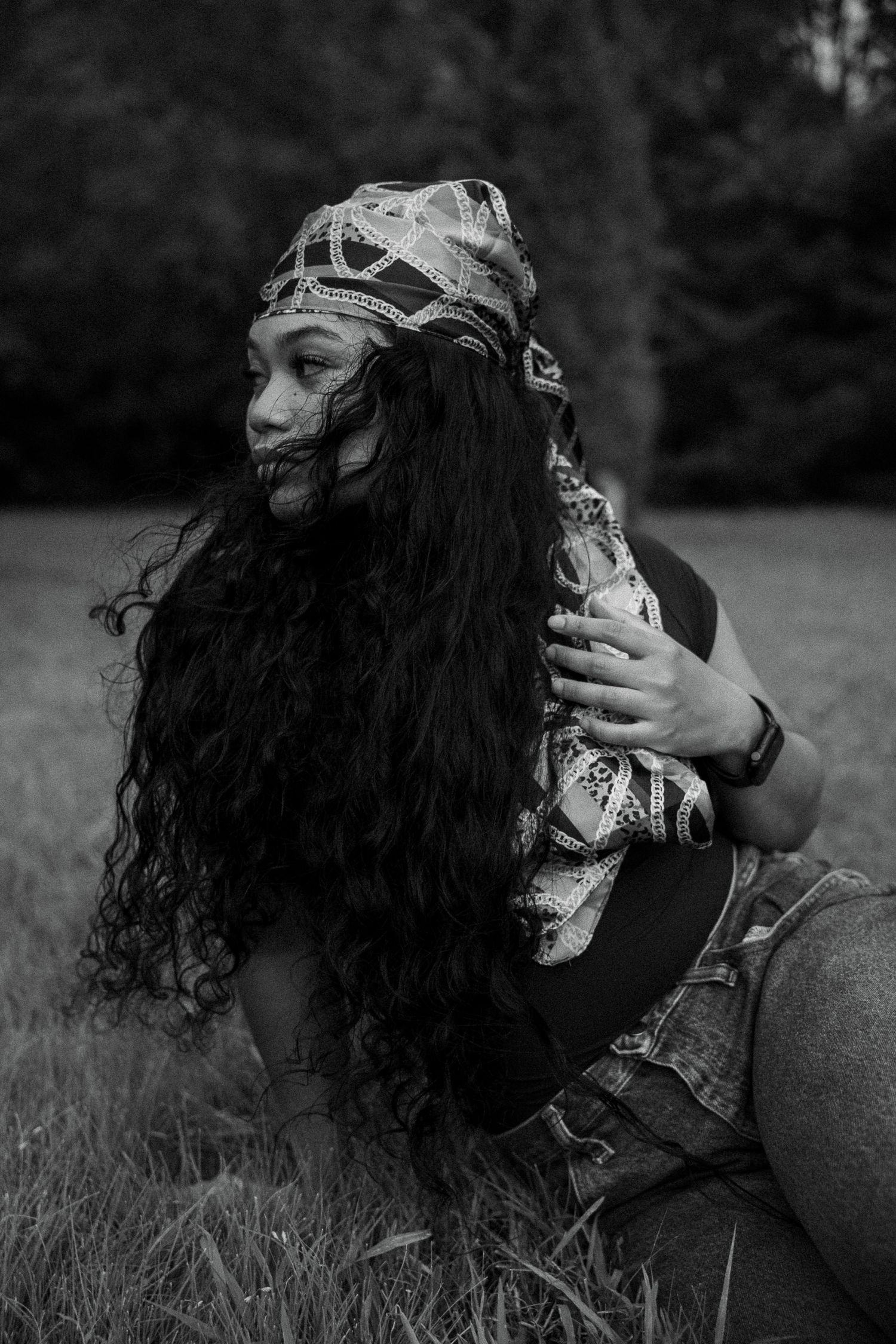 Black and white photo of woman with long curly hair and patterned headscarf seated in grassy field.