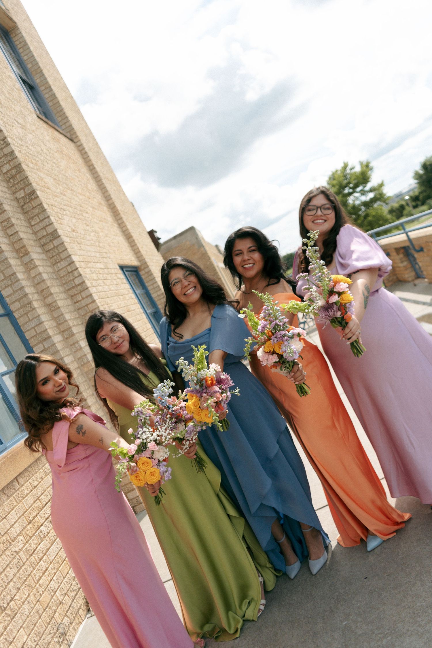 Five bridesmaids in colorful gowns holding floral bouquets pose outdoors at a wedding celebration.