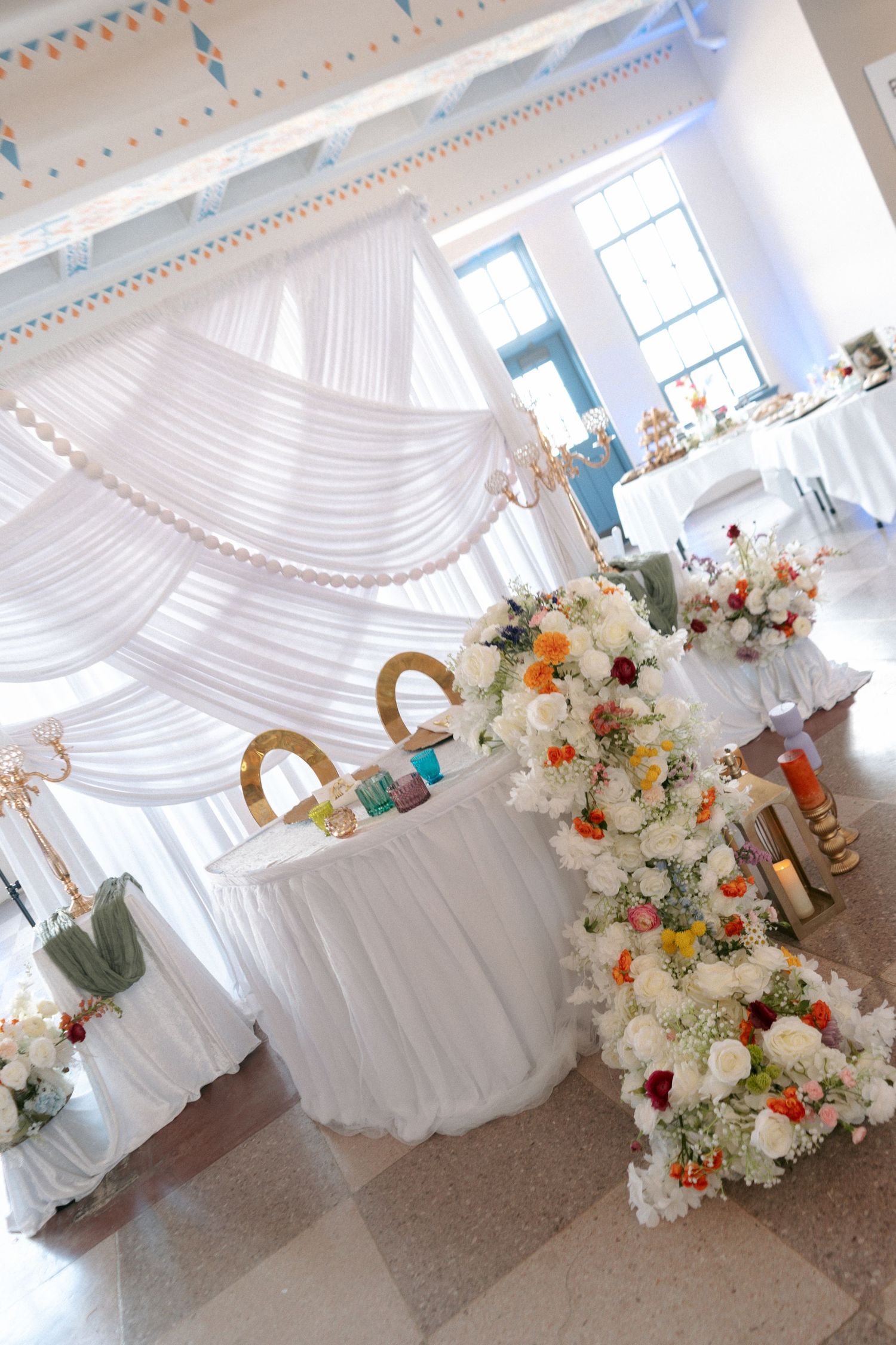 Elegant wedding reception venue with white draped canopy, floral arrangements featuring white, orange, and red blooms on decorated tables.