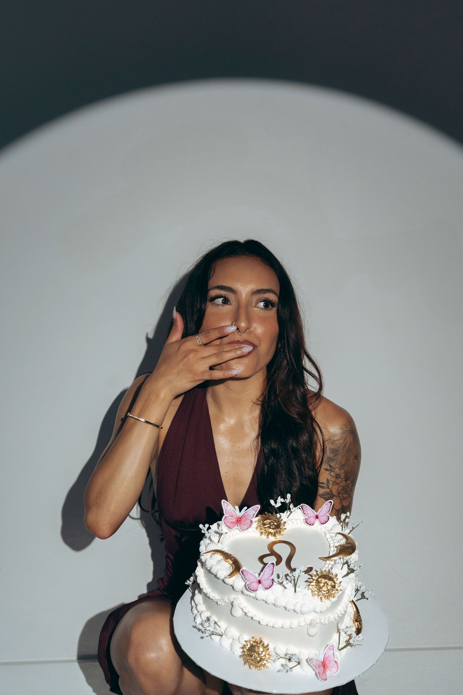 Woman in burgundy dress holding decorated white birthday cake with butterflies and gold accents, hand over mouth.