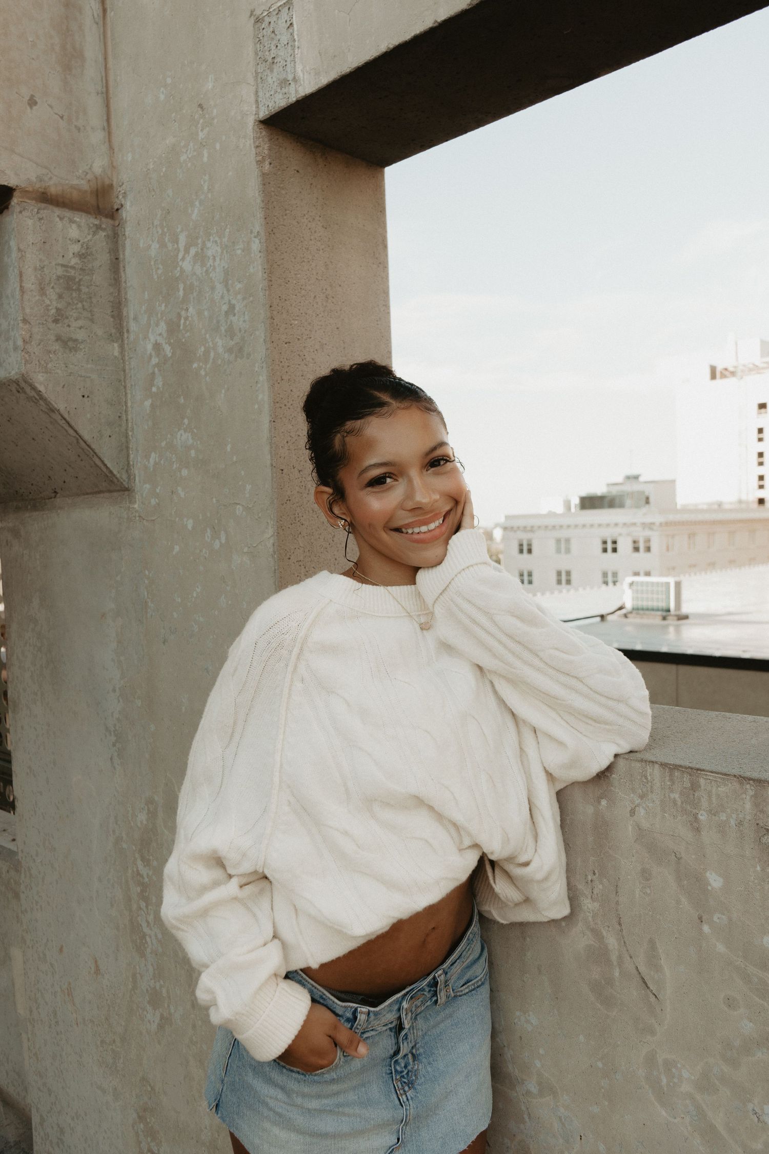 A cheerful young woman in a white oversized sweater and denim skirt poses on an urban rooftop.