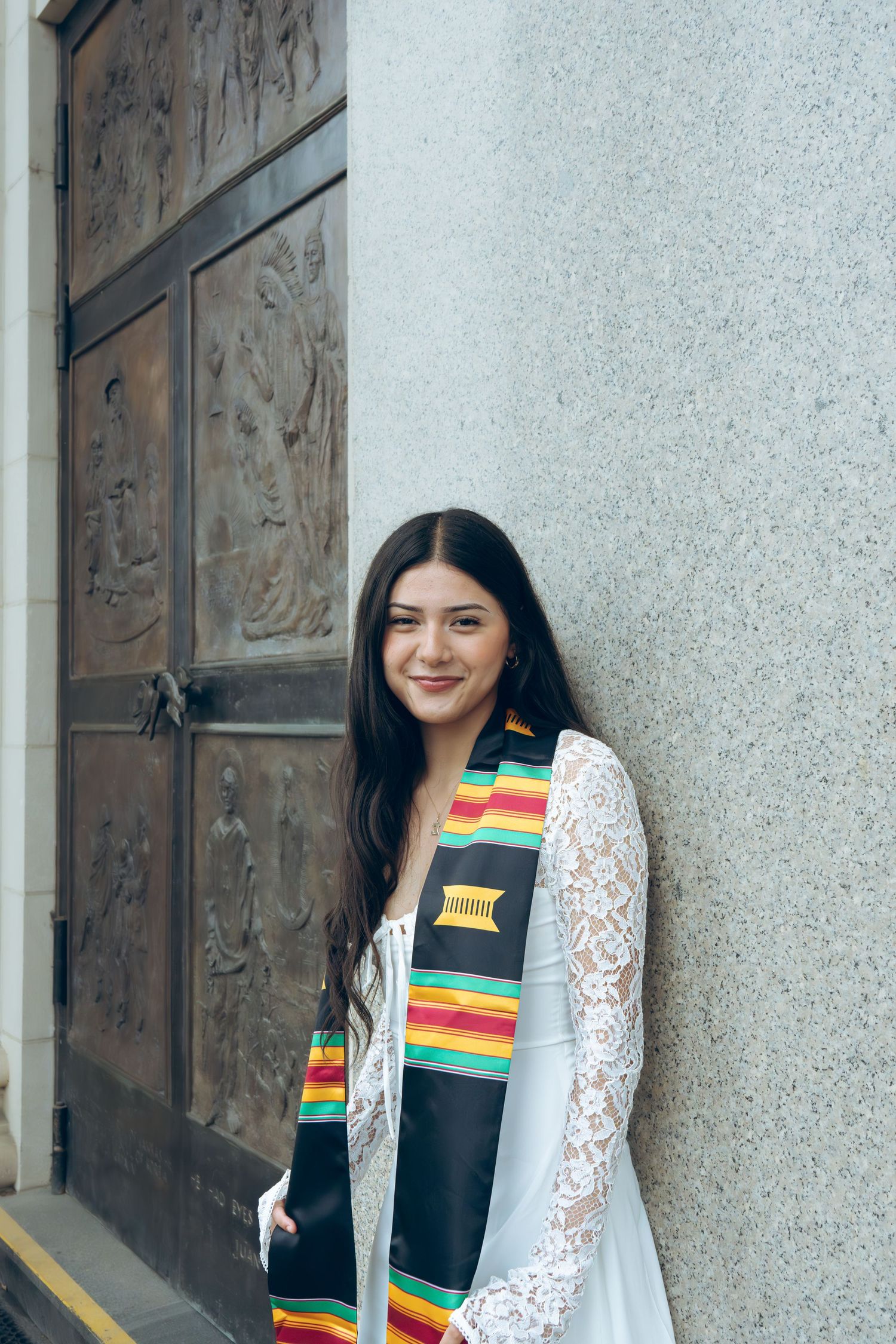 Young woman in white lace dress with colorful graduation stole posing by ornate door.