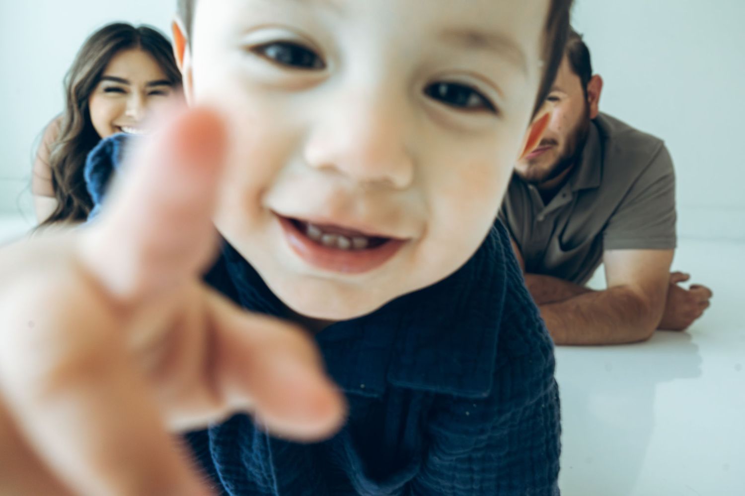 Adorable toddler boy reaching toward camera while parents smile in background.