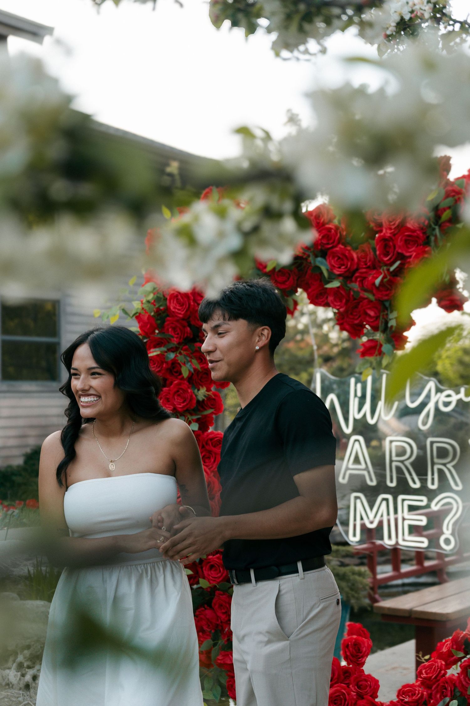 Couple smiling together under red rose heart arch with Will You Marry Me neon sign proposal setup.