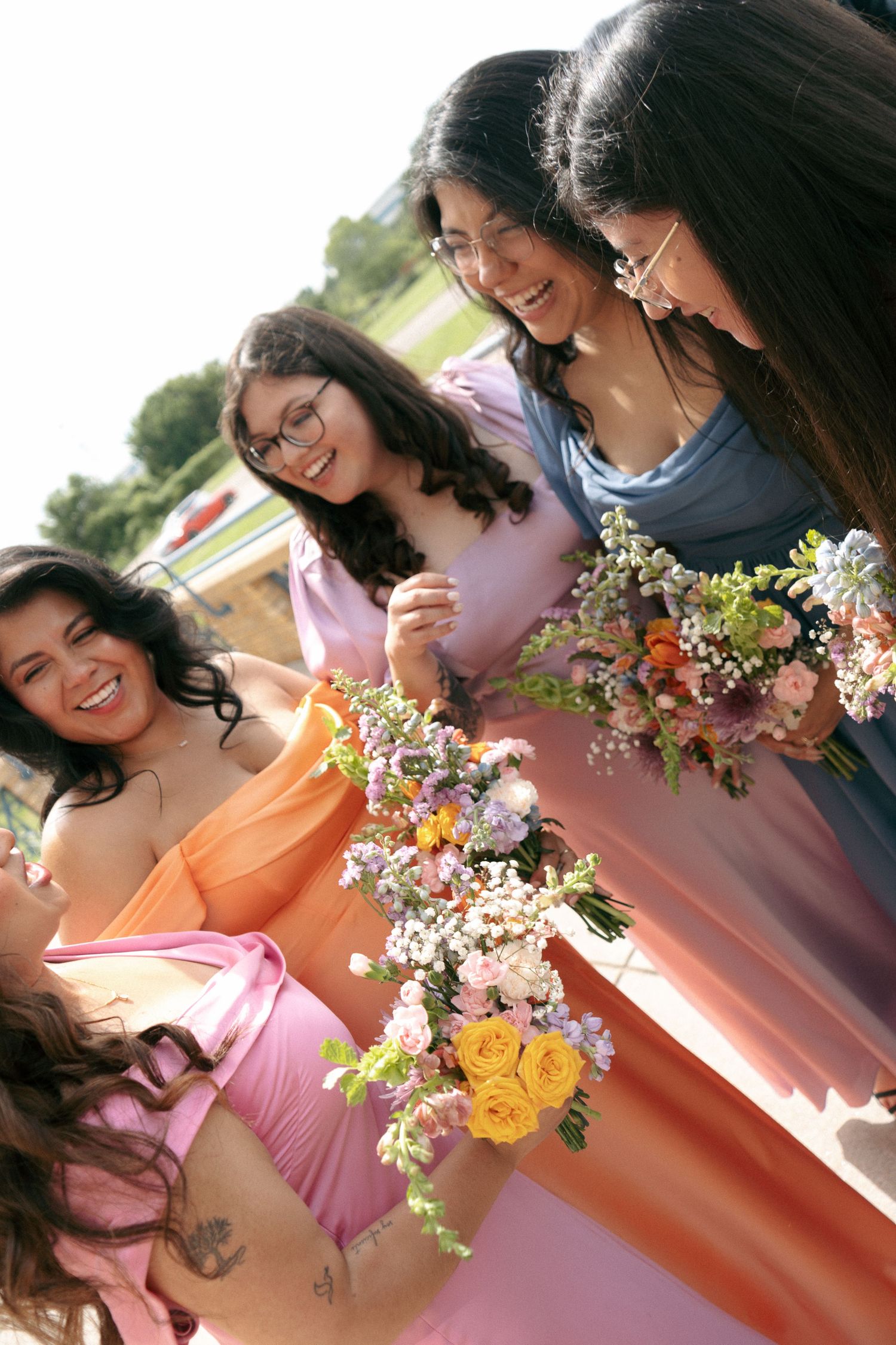 Bridesmaids in pink, orange, and blue dresses laughing together while holding colorful floral bouquets at an outdoor wedding.