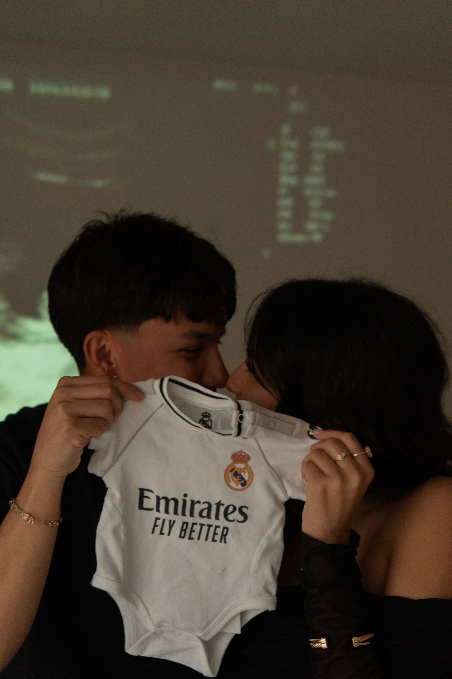 Couple kissing behind a Real Madrid Emirates baby onesie in front of an ultrasound projection.