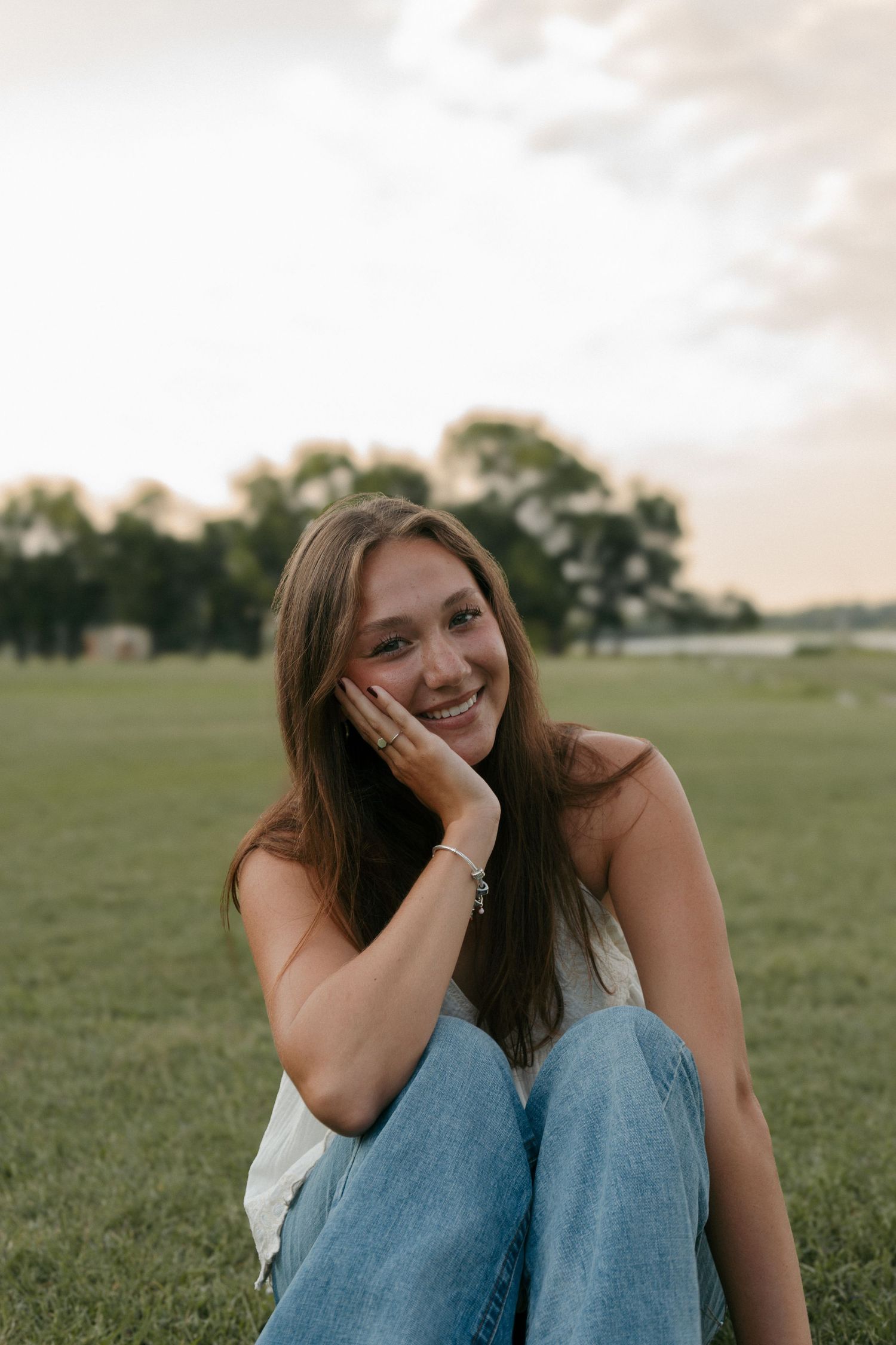Young woman with long brown hair sits on grass, smiling, wearing white top and blue jeans at sunset.
