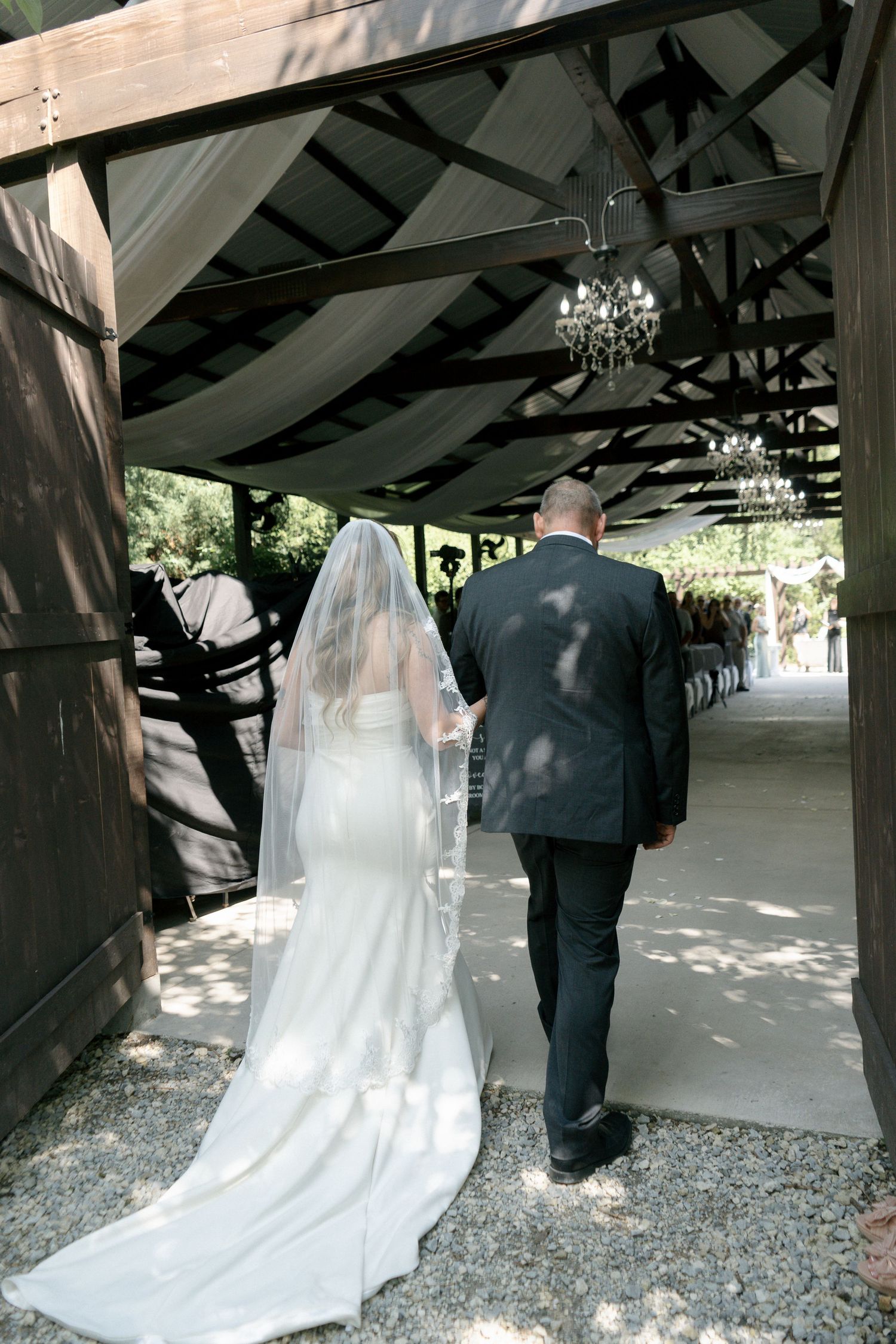 Bride in white gown with long veil walks with escort down aisle under chandelier-lit wooden pergola at wedding.