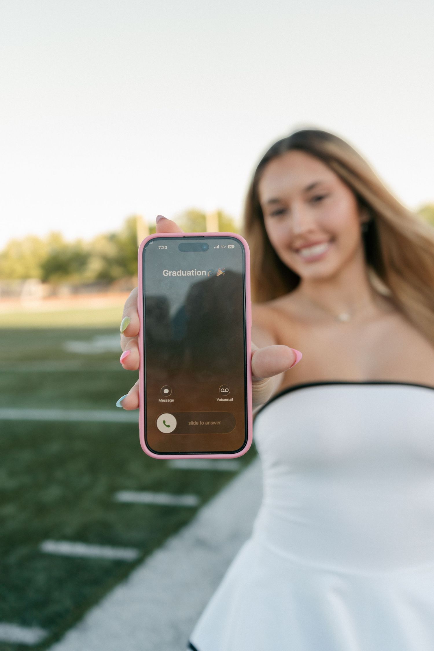 Smiling young woman in white dress holding pink iPhone showing Graduation call screen on football field.