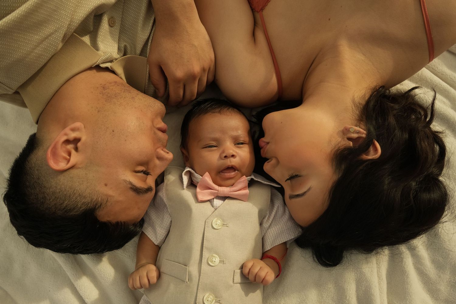 Happy parents lying with their newborn baby between them on a white bed.