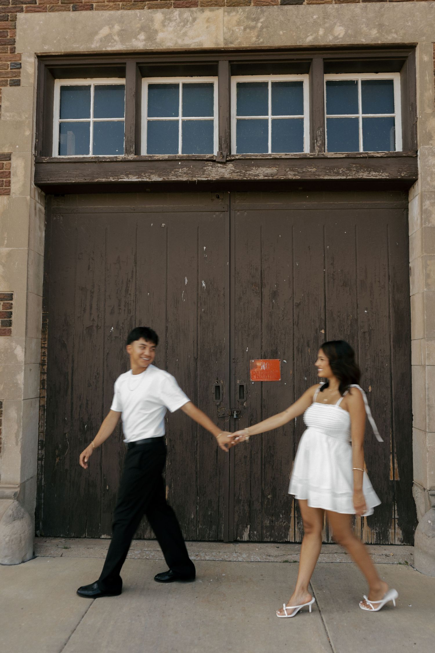 A young couple in formal attire holds hands while walking in front of a large wooden door on a brick building.