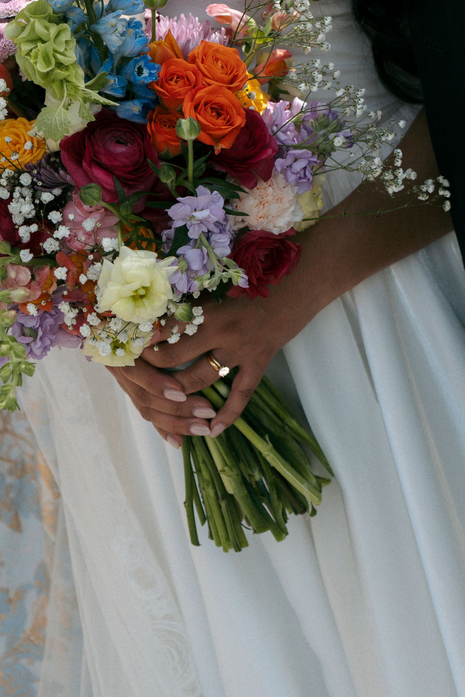 Bride holding a vibrant wedding bouquet with orange, red, purple, and white flowers in a white gown with a gold ring.