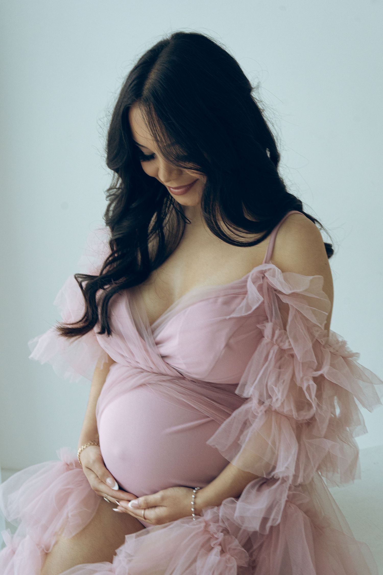 Smiling pregnant woman in pink tulle off-shoulder maternity gown gently holds baby bump in bright studio.