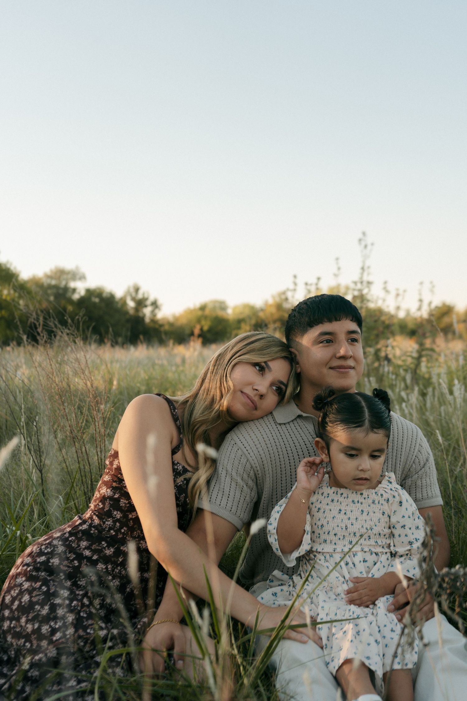 A family of three sits peacefully in tall grass at sunset, with the mother leaning on the fathers shoulder.
