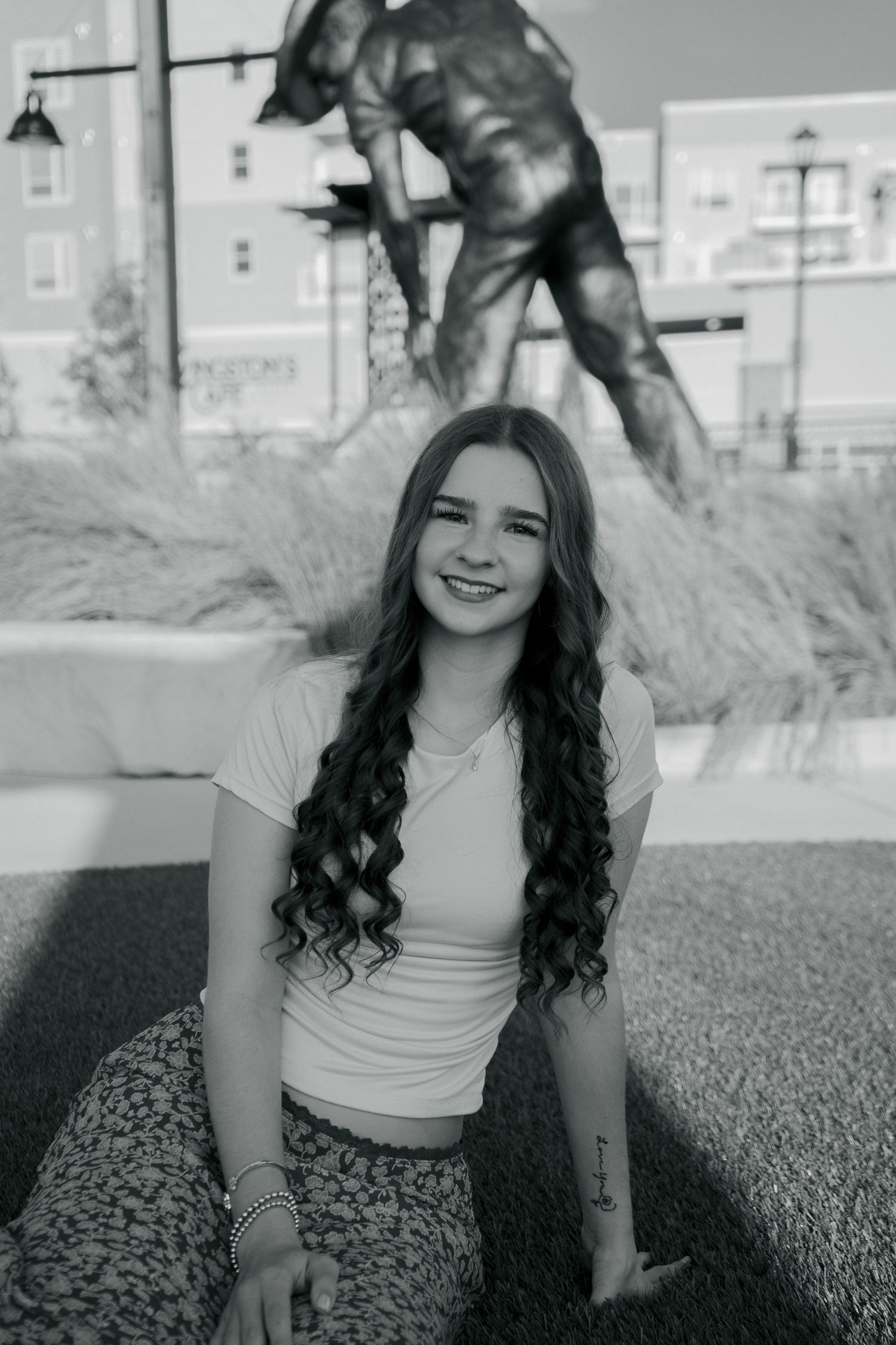 Black and white portrait of smiling teenage girl with curly hair sitting outdoors near bronze statue.