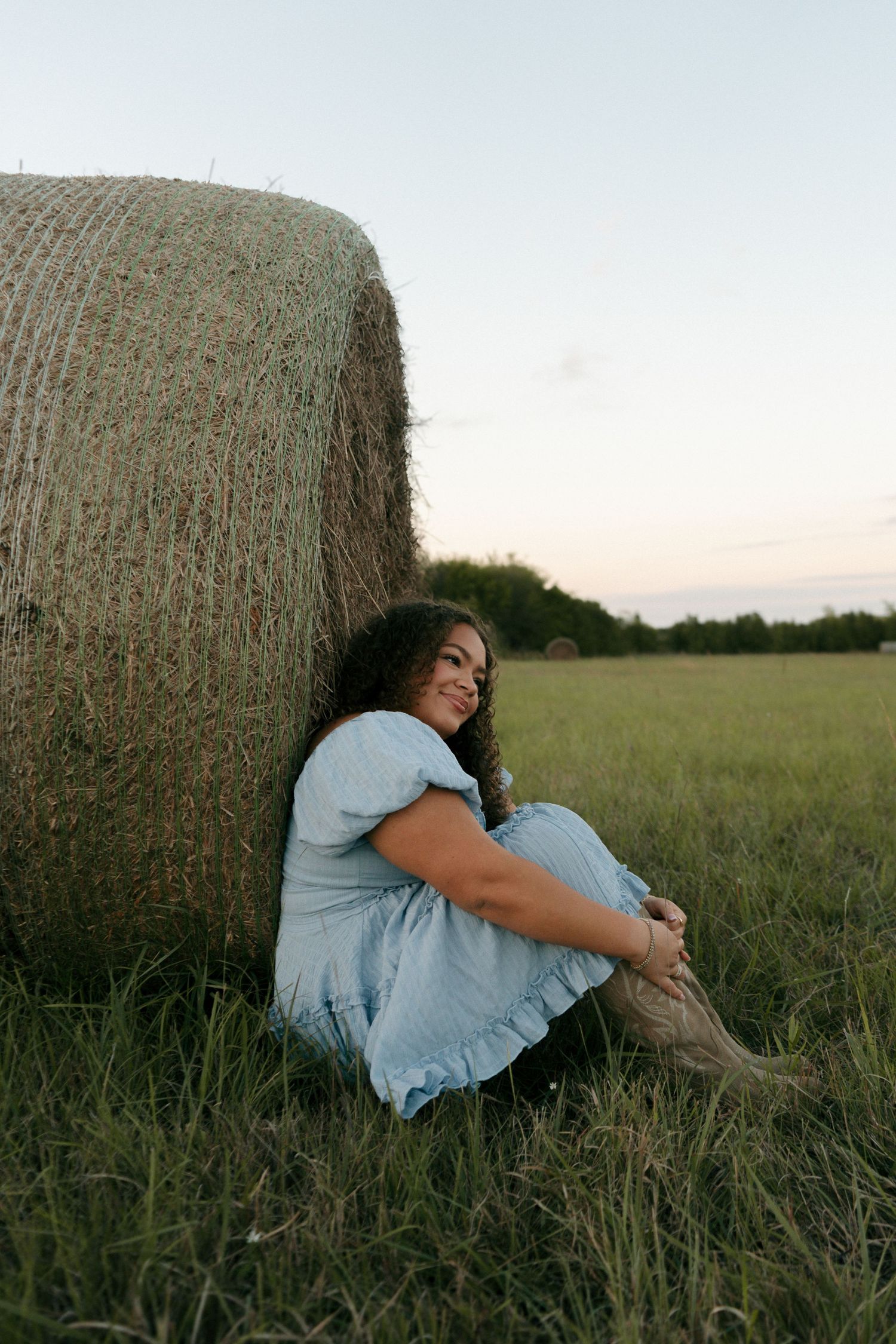 Young woman in blue dress and cowboy boots sitting and smiling beside a hay bale at sunset.