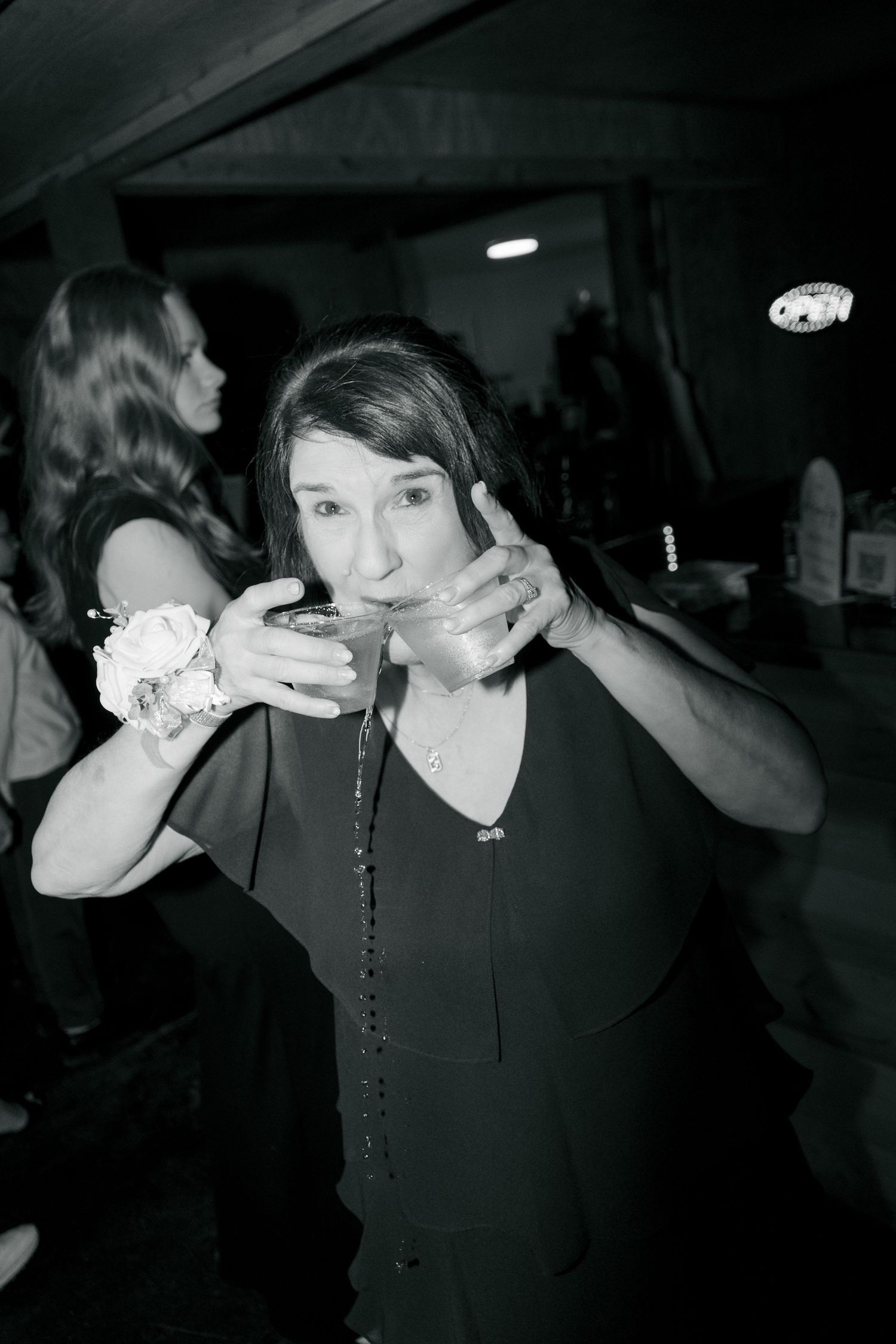 Black and white photo of a woman drinking from a cup at a party, wearing a corsage and necklace.