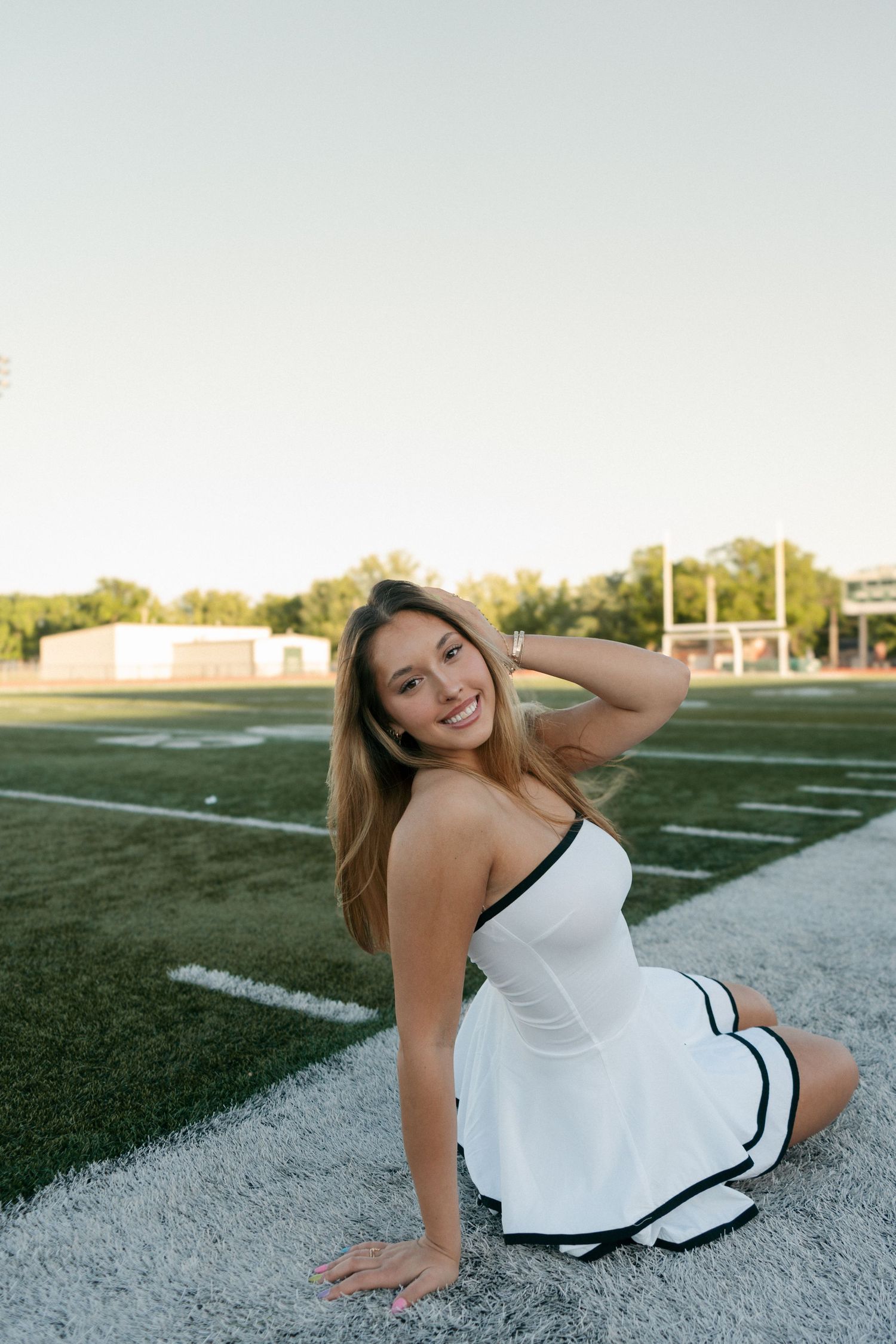 Young woman in white strapless dress sitting on football field sideline smiling at camera.