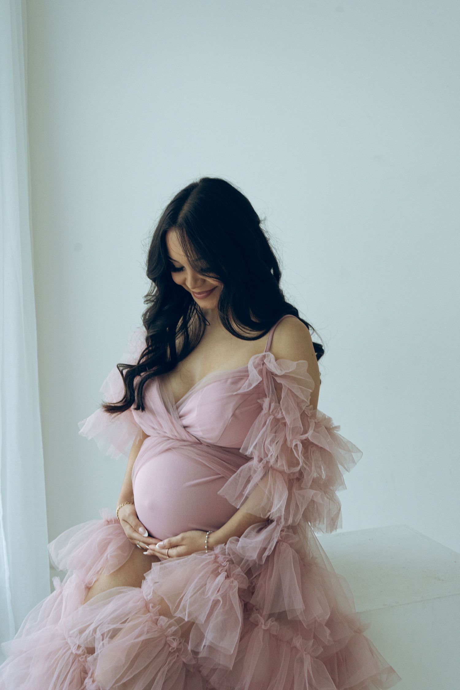 Pregnant woman in elegant pink ruffled tulle maternity dress cradles baby bump during studio photoshoot.