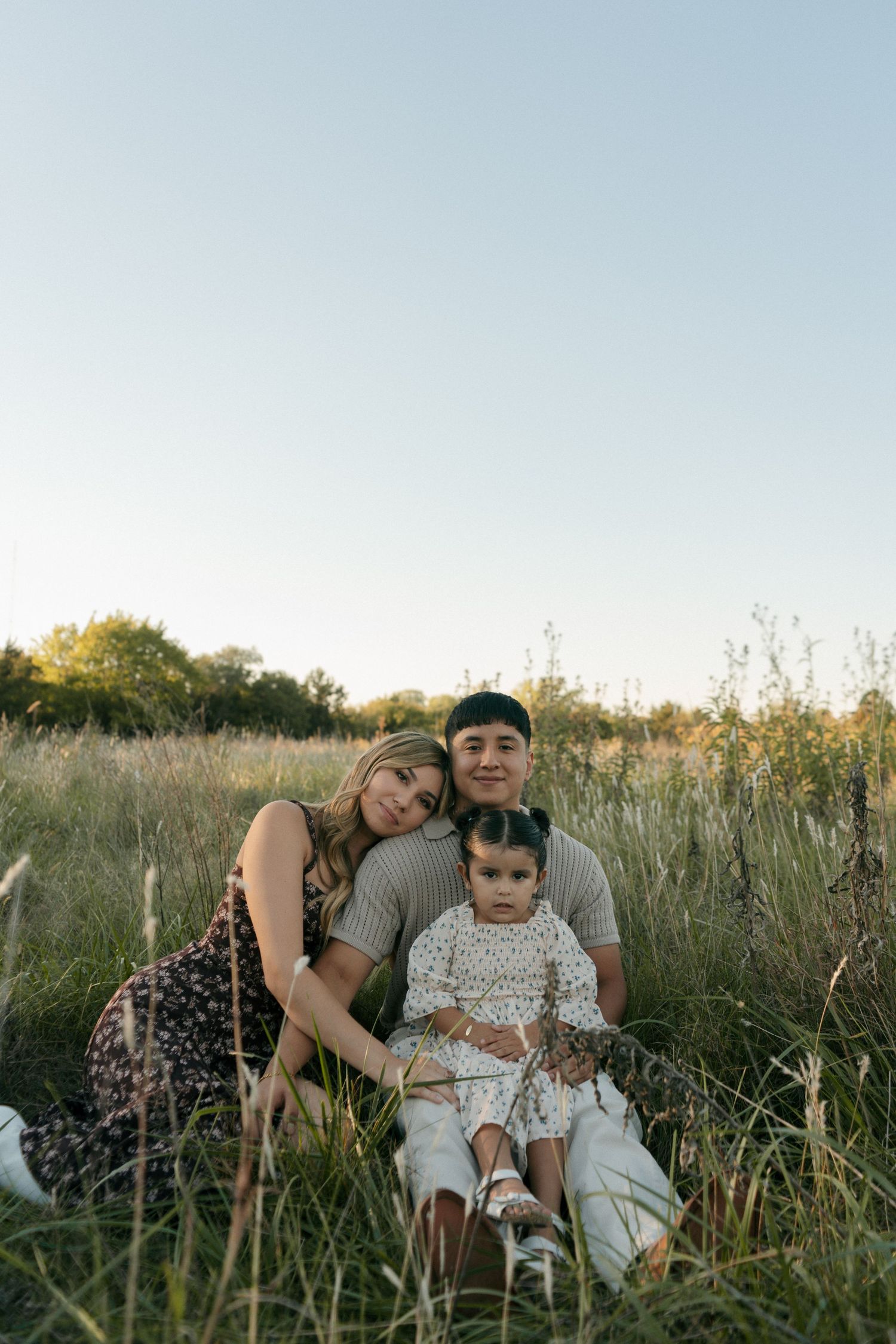 A happy family of three sits together in a lush green field, smiling at the camera during golden hour.