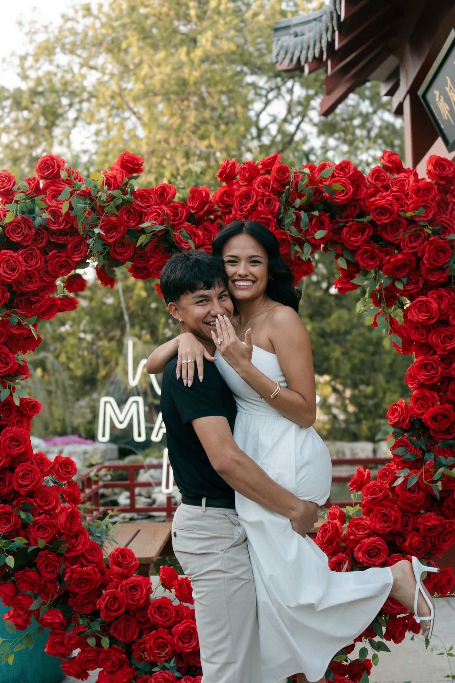 Happy couple embraces under a red rose arch during a romantic outdoor engagement proposal celebration.