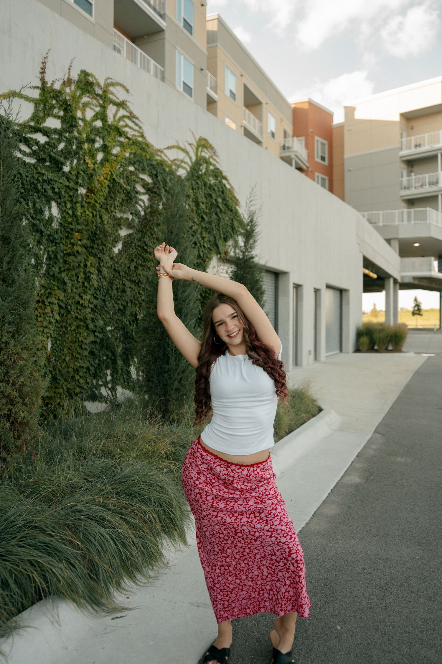 Smiling young woman in white tank top and red floral maxi skirt dancing outside near green ivy wall.
