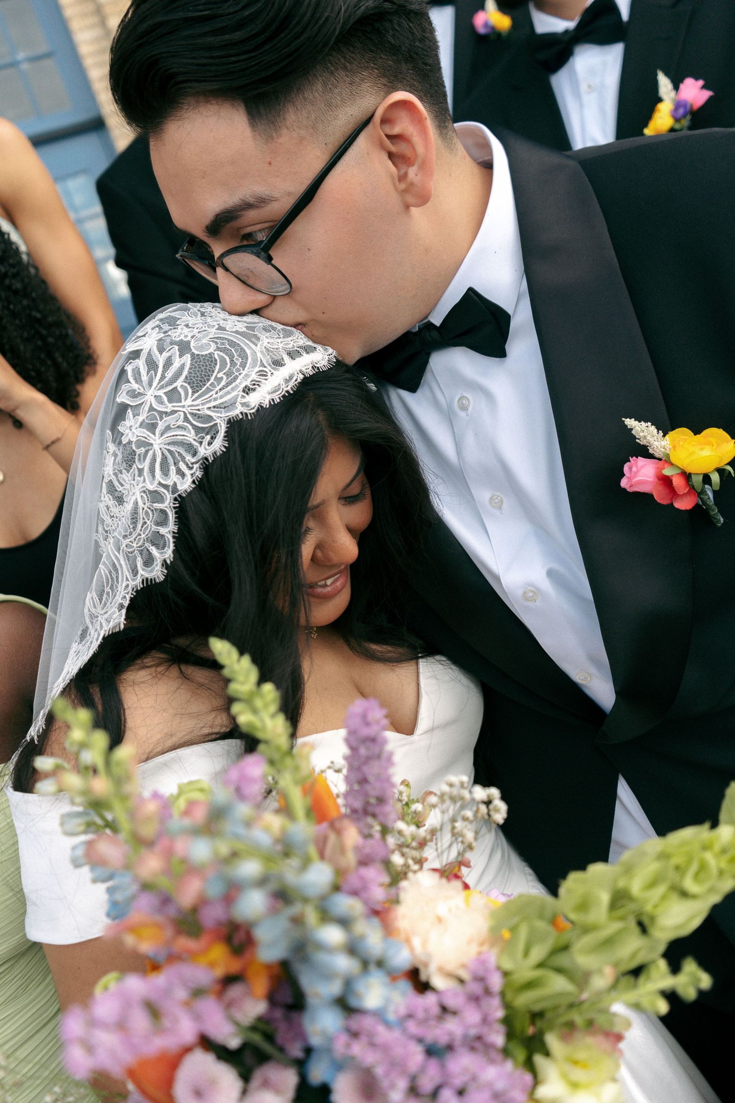 Groom in black tuxedo kisses crying bride in white dress and lace veil while holding colorful wedding bouquet.
