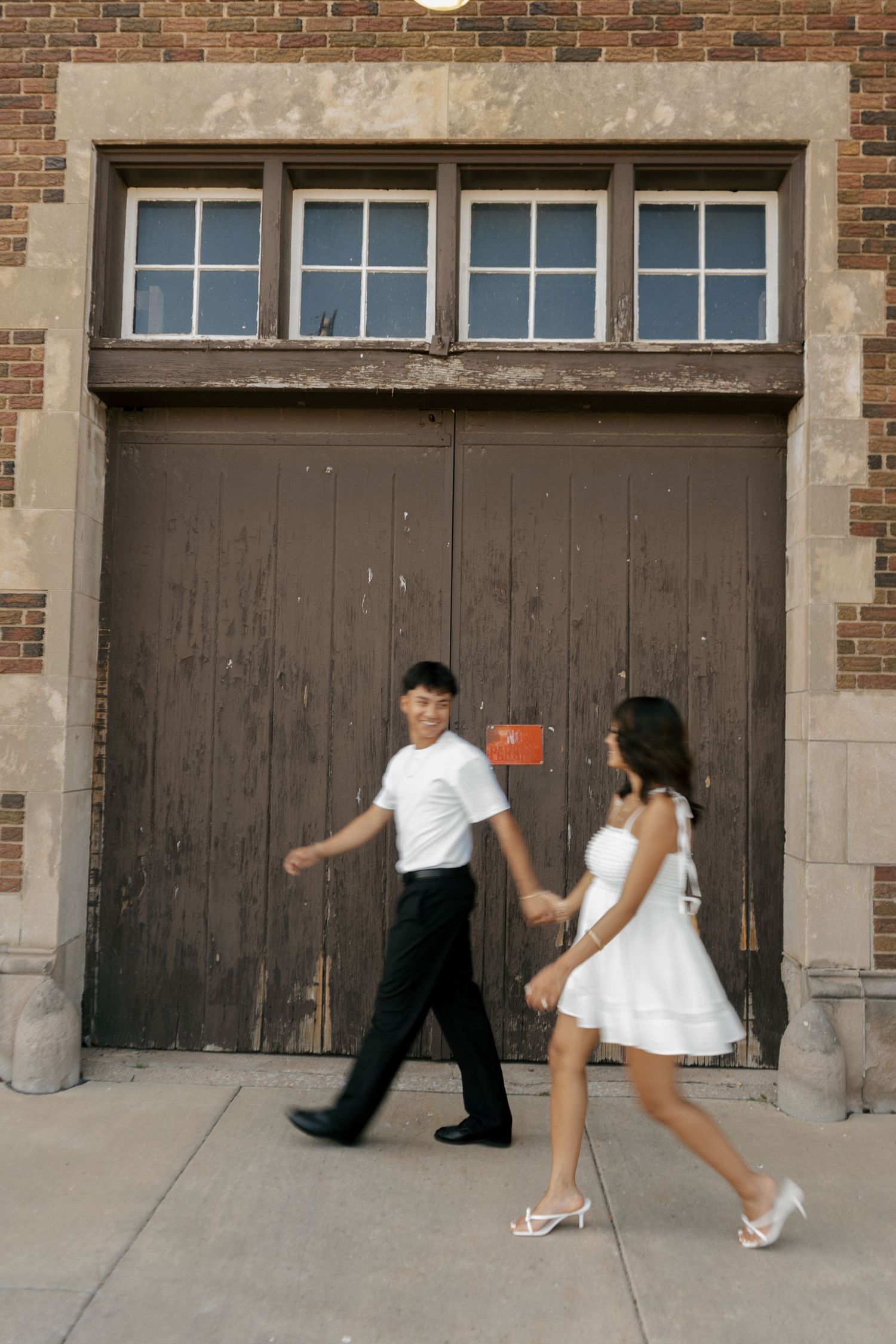 A couple in formal attire holds hands while walking past a large wooden door on a brick building.