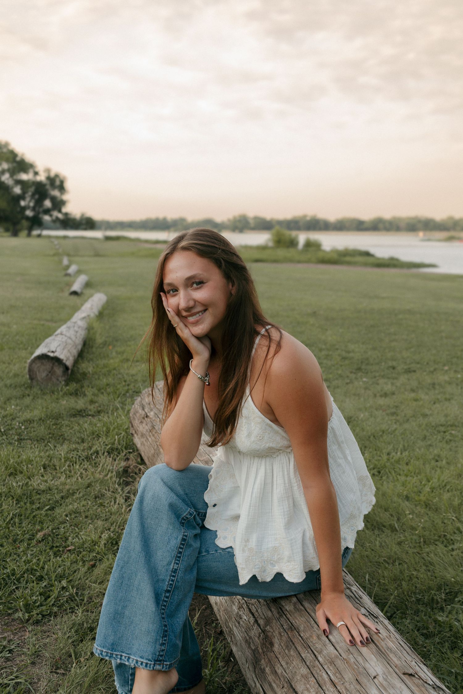 Smiling young woman in white lace top and jeans sits on a log near a lake at golden hour.