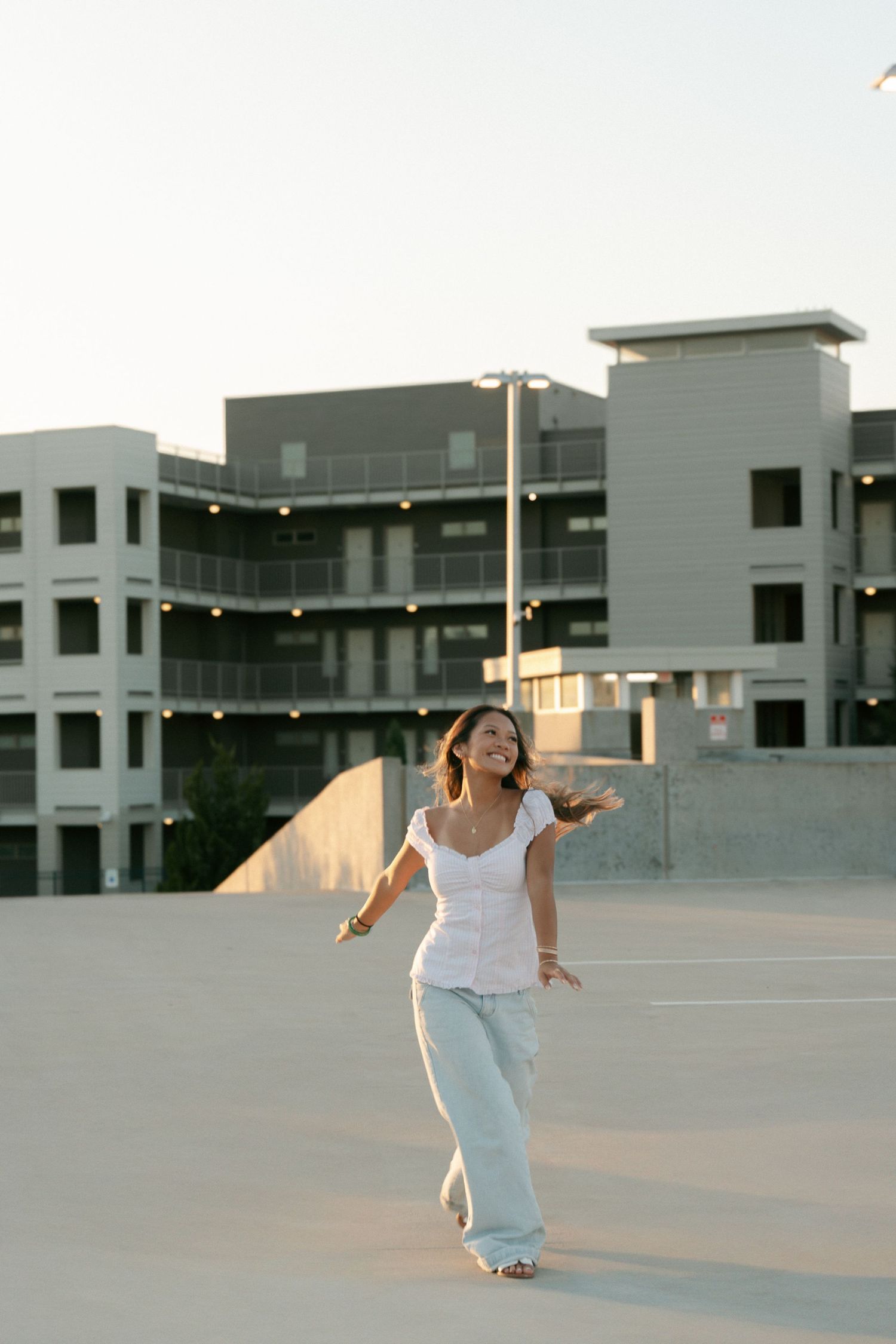 Joyful young woman in white top and jeans laughing on rooftop parking lot at golden hour.