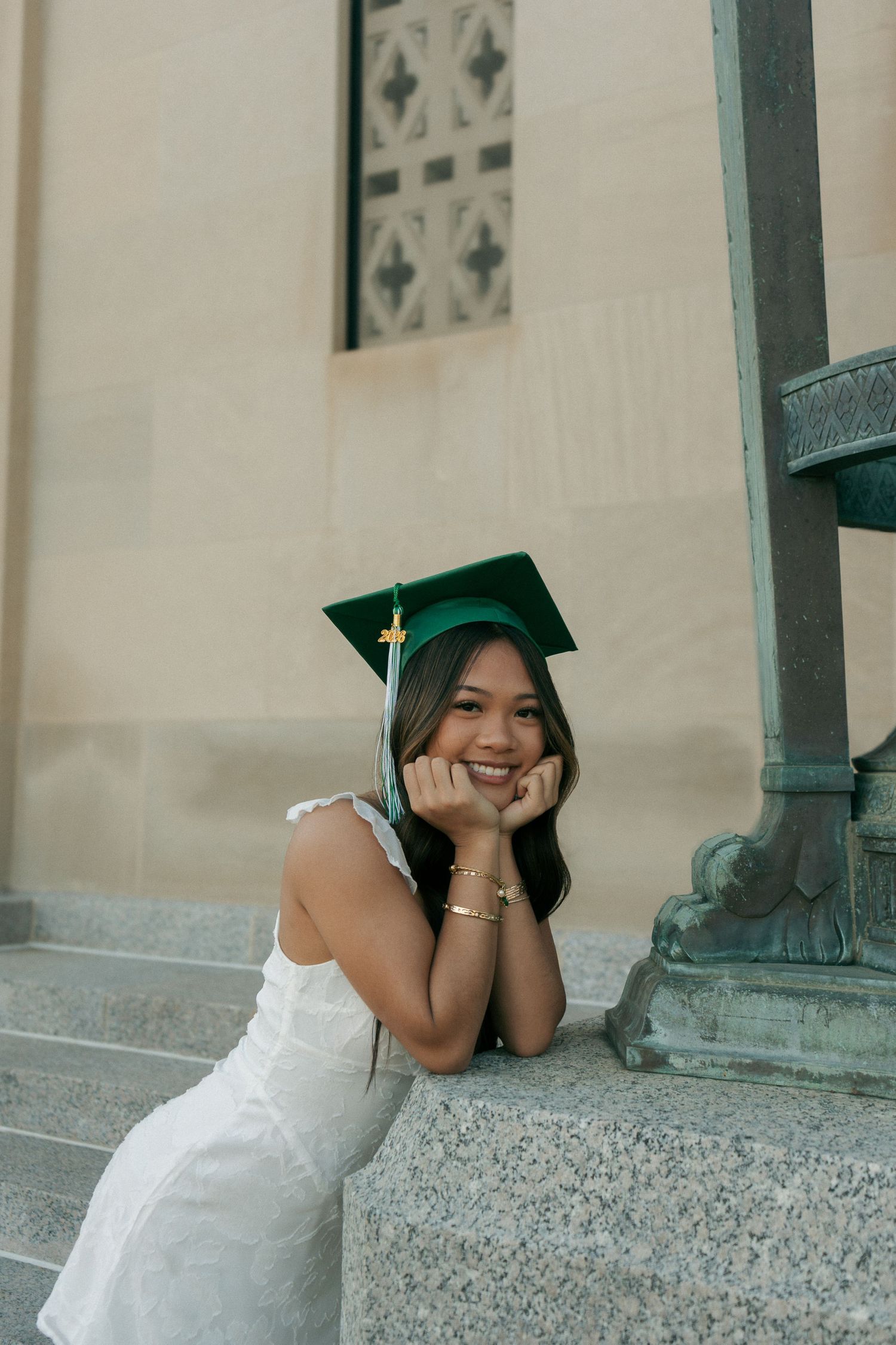 Happy graduate wearing green cap and white dress posing on stone steps for senior portrait photos.